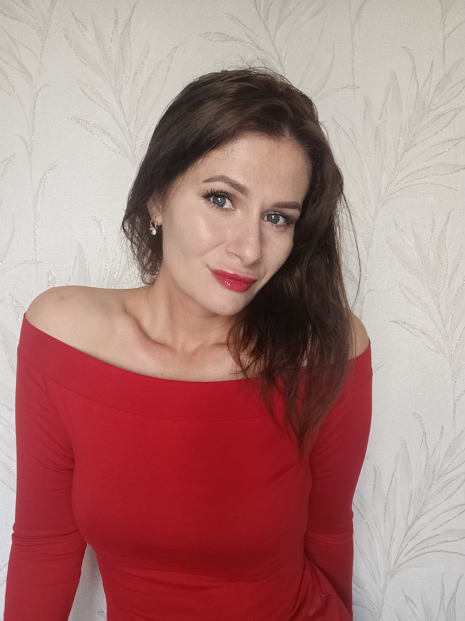 The image features an adult female centered in an indoor portrait. She has dark brown, wavy hair that falls over her shoulders. Her eyes are blue, and her lips are made up with glossy red lipstick. She wears a red, off-the-shoulder, long-sleeved top or dress. A small gold-toned hoop earring with a white pearl-like bead is visible in her left ear. Her head is tilted slightly, and she looks directly at the viewer with a subtle smile. The background is a light-colored wall covered in wallpaper featuring a repeating pattern of stylized, silvery-grey botanical or feather-like motifs with a slight sheen. The location is an indoor setting; no discernible city or specific geographical information can be determined, rendering the location as "Unknown, Unknown."