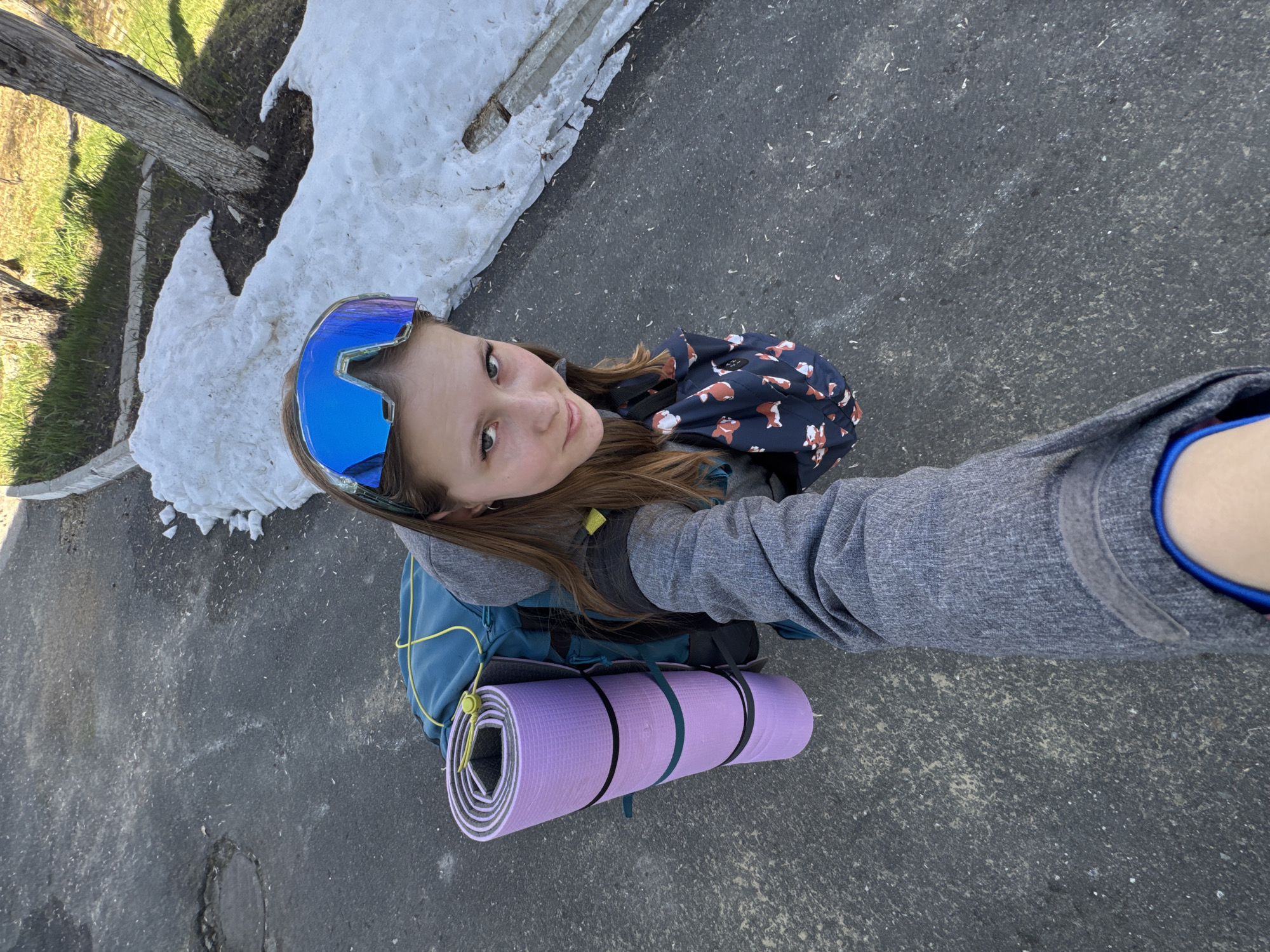 A young person, likely female, takes a selfie outdoors on a gray paved surface, possibly a road or sidewalk. The subject is wearing mirrored blue ski goggles, a gray hooded sweatshirt, and a backpack. A rolled-up purple sleeping mat is strapped to the backpack. A fox-printed backpack or bag is visible on her back. Patches of melting snow are present to the side near a tree, suggesting the location might be in a colder climate. Based on the request, the location is suggested as Berezniki, Russia.