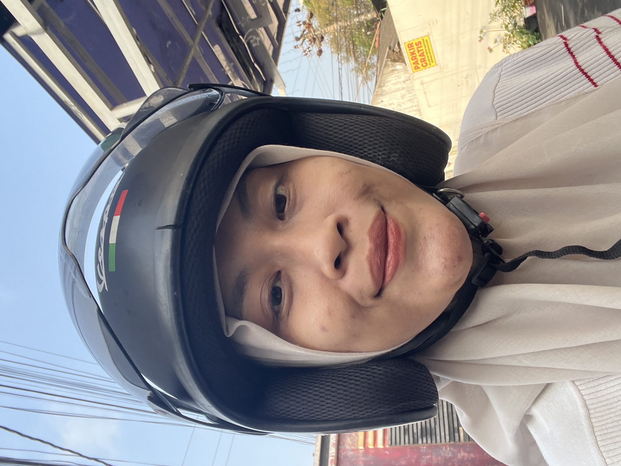 A selfie shows a young Indonesian woman wearing a black helmet and a light-colored hijab. She is outdoors, as indicated by the clear blue sky and power lines visible in the background. The woman has fair skin, light makeup, and is smiling slightly at the camera. She is wearing a light-colored sweater with red stripes. In the background, there's a building with a blue roof structure and a yellow sign that reads "PARKIR GRATIS" (Free Parking in Indonesian), suggesting a public area or business in Kecamatan Pati, Indonesia. The overall lighting suggests it is daytime.