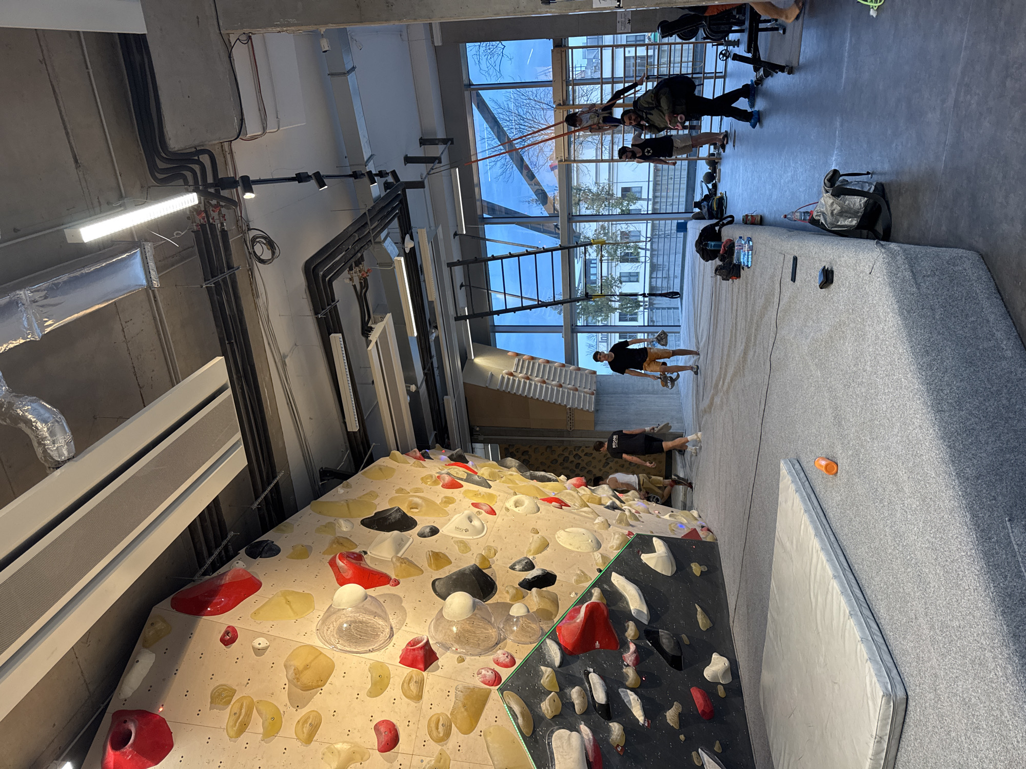 The image shows an indoor climbing gym in Sofia, Bulgaria. The foreground features a large bouldering wall with various colored climbing holds. To the right of the bouldering wall, there is a crash pad. In the background, there are windows through which one can see buildings and trees. Several people are engaged in various activities. Some individuals are climbing, while others appear to be stretching or exercising in the background. Bags and personal items are scattered on the floor. Overhead, there are industrial-style lighting fixtures, ventilation ducts, and exposed pipes.