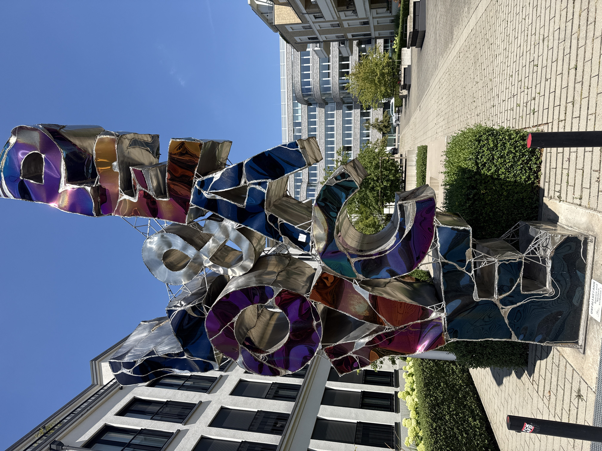 The image shows a large, abstract metal sculpture in a public outdoor space. The sculpture is composed of fragmented, geometric shapes that are reflective and appear to be made of iridescent metal, displaying a spectrum of colors including blues, purples, and oranges. The letters "YO", "CA", and possibly parts of other letters are discernible within the sculpture.
The sculpture is situated in front of modern white buildings with black-framed windows and balconies. To the right of the sculpture, a paved walkway stretches into the distance, flanked by more contemporary buildings with brick and white facades. Trees and neatly trimmed green bushes are interspersed throughout the scene. The sky is clear and blue, indicating a sunny day.
A sign is visible at the base of the sculpture which reads "UNFALLGEFAHR Rutschgefahr verbote" (Danger of accident, risk of slipping, prohibited). Another black bollard with a red top is in the foreground to the left.
This sculpture is located in front of the Commerzbank building in Dusseldorf, Germany, at the address Schadowstraße 48-50.