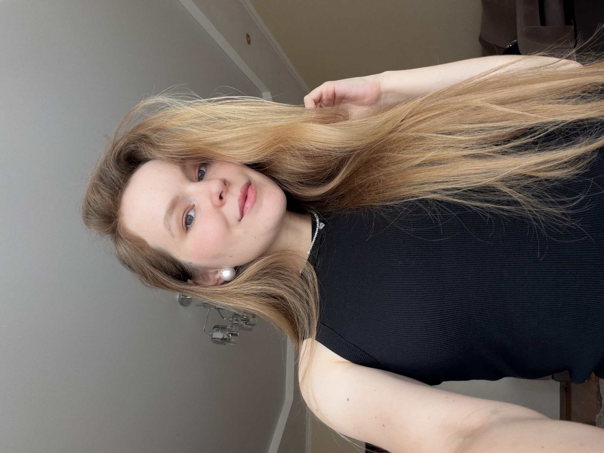 A young woman with long, blonde hair is shown in a selfie. She is wearing a black ribbed tank top and a pearl earring. The background appears to be an indoor setting, possibly a living room, with a chandelier visible on the ceiling. The woman is looking directly at the camera with a slight smile. There is no indication of the city of Kazan, Russia.