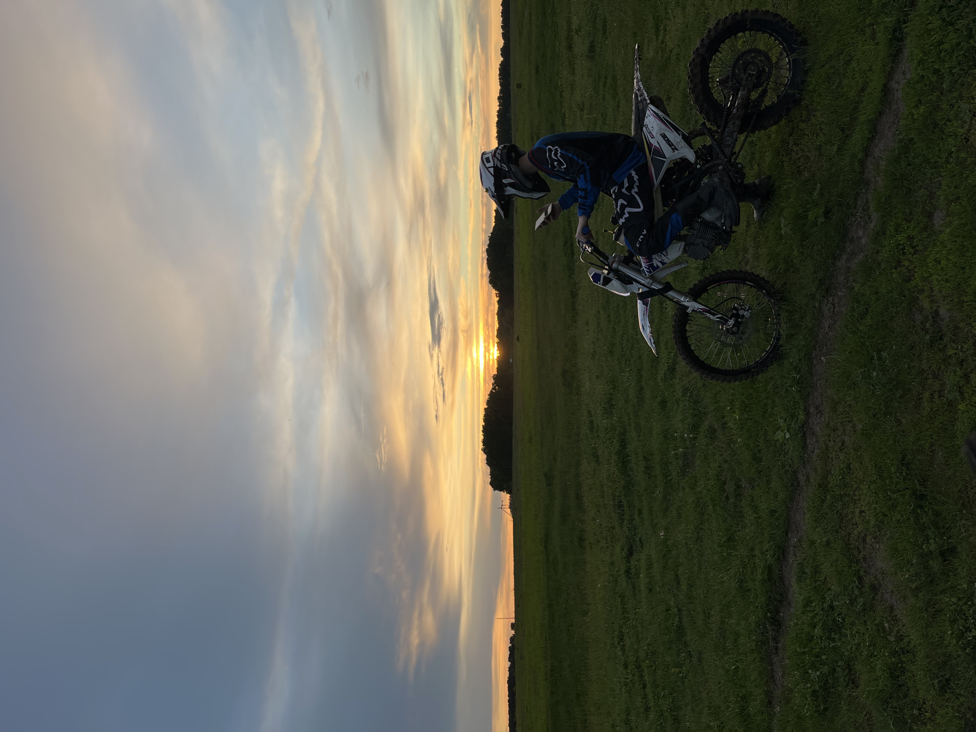 A medium, outdoor shot depicts a man on a dirt bike against a colorful sunset. The location is an open, grassy field. The man, wearing a helmet and riding gear, is partially visible and appears to be interacting with the bike. The dirt bike is a prominent object. The sunset is a key environmental detail, coloring the sky with shades of orange and grey. The ground shows signs of tire tracks in the grass. The location cannot be determined to be Новочувашский, Russia.