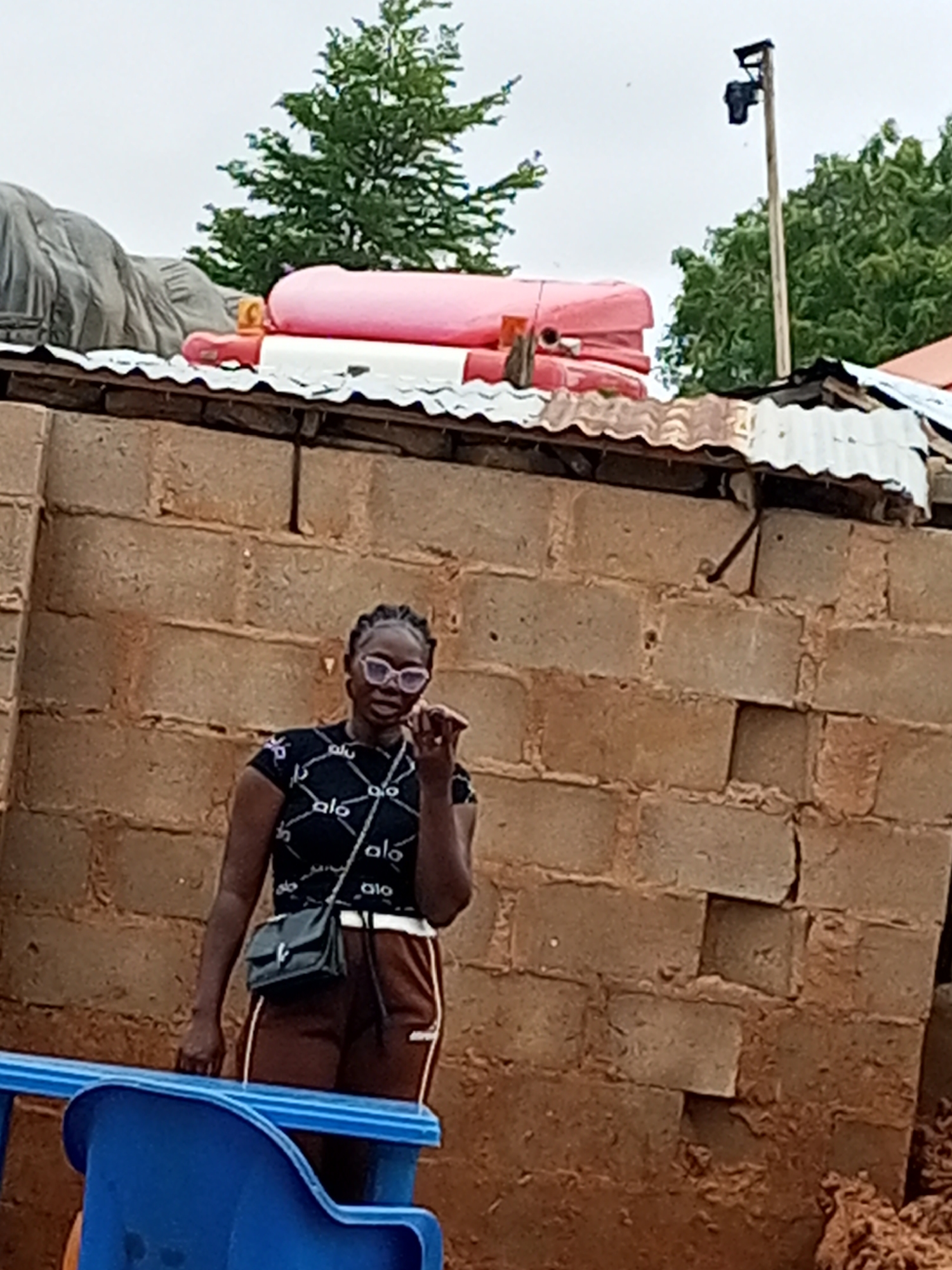 A young woman with dark braided hair and pink-rimmed glasses stands in front of a brick wall in Tempure, Nigeria. She wears a black t-shirt with a white "alo" pattern and brown athletic pants with white stripes. A dark purse with a strap is slung across her body. Her right hand is raised, touching her chin. In the background, a corrugated metal roof is visible, topped with large pink and red objects, possibly inflatable toys. Trees and a utility pole with a camera are also present in the background. A blue plastic chair is in the foreground.