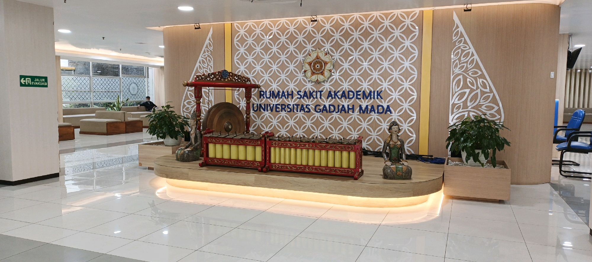 The image depicts the lobby of a building, identified by the text "RUMAH SAKIT AKADEMIK UNIVERSITAS GADJAH MADA" on a wall. The lobby features a polished tile floor with a reflective surface. In the center, a wooden stage is set up, adorned with traditional Indonesian musical instruments, including a large gong and a gamelan. A statue of a seated figure is placed to the right of the instruments. The backdrop is a wall with a repeating geometric pattern and the university's emblem. To the left, there are seating areas with sofas and wooden-topped tables. An "EVACUATION ROUTE" sign is visible on the wall to the far left. A single person is seated on a sofa in the background, wearing a mask. Several potted plants are placed around the lobby, adding greenery. In the background on the right, blue chairs are visible. The lighting in the scene appears to be from overhead fixtures and accent lighting under the stage. The overall impression is a clean, modern, and culturally decorated interior space. The location is specified as Trihanggo, Indonesia.
