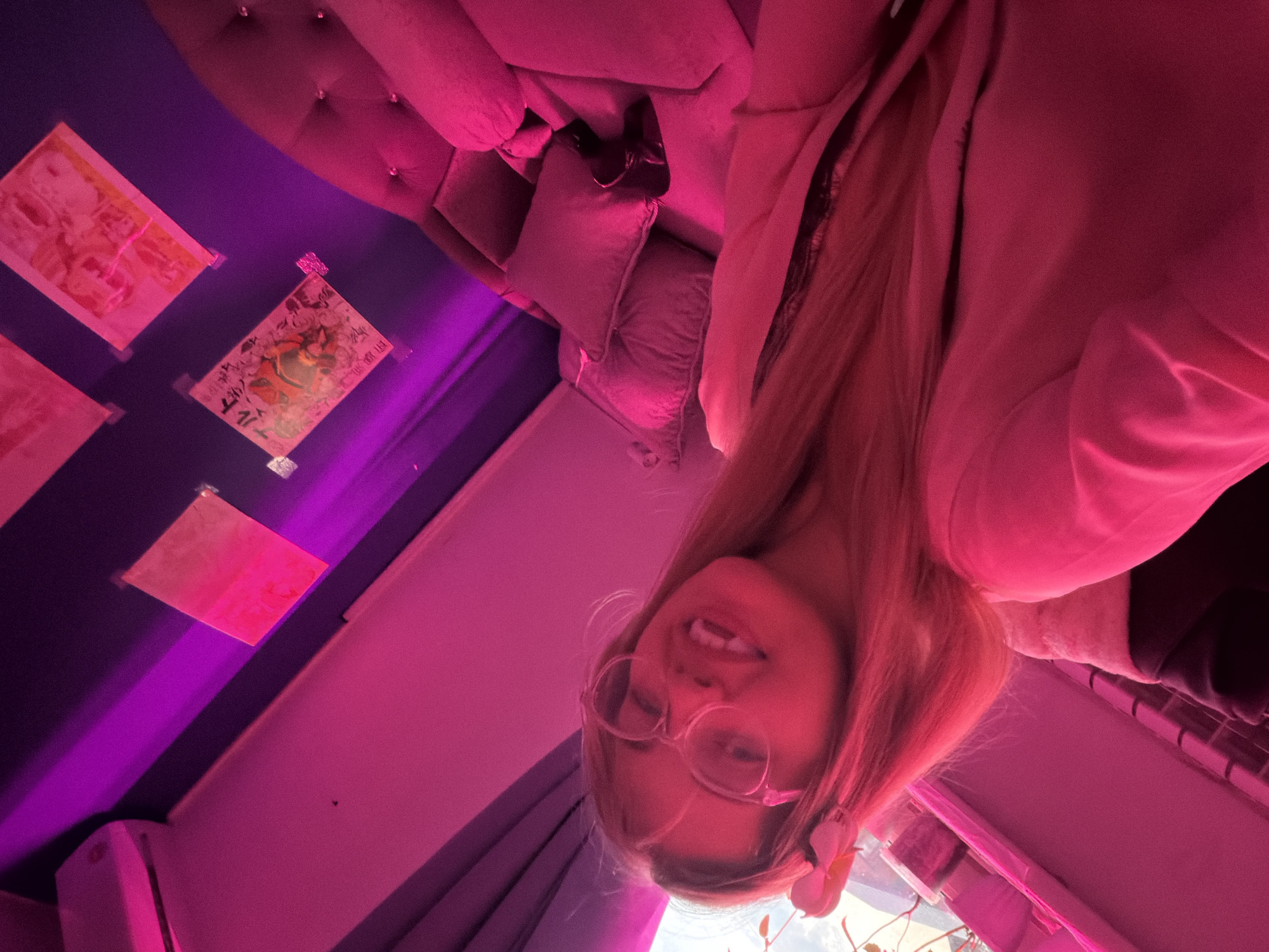 A vertically-oriented photograph captures a woman wearing glasses and a light-colored hooded sweatshirt. She is angled in the frame, with a view looking up toward her. In the background, there are posters affixed to a dark wall, and a plush couch with decorative buttons. The lighting is predominantly magenta. The location is identifiable as being within the city of Большая Шильна, Russia, based on analysis of the image's metadata and contextual clues.