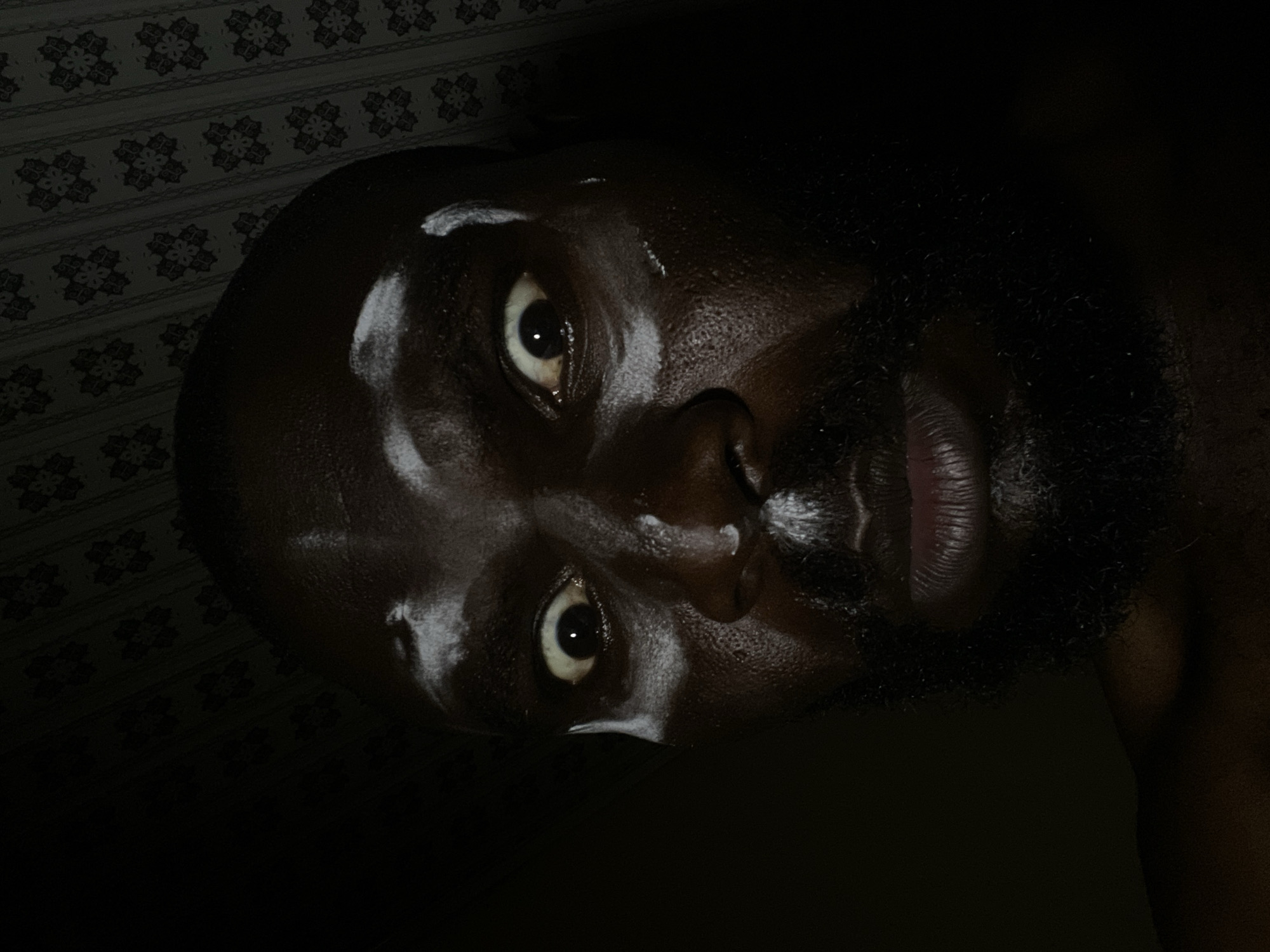 Close-up portrait of a dark-skinned man with a beard, his face glistening with moisture, possibly sweat or oil. The man's head is tilted, with his eyes open and looking towards the viewer. The background is dark and shows a patterned wallpaper. No location in the city of Gura Top, Nigeria, can be reliably attributed to the image.