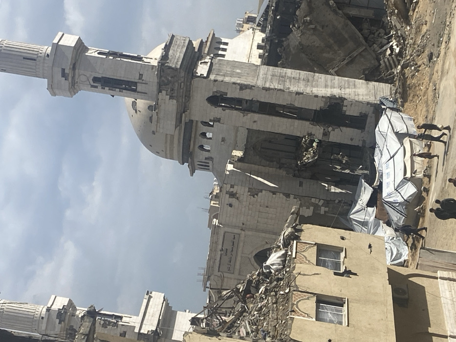 The image depicts a scene of extensive destruction centered around the Grand Al-Omari Mosque in Khan Yunis, Palestinian Territory. The mosque's main structure, identifiable by its dome and two minarets, is severely damaged. The dome exhibits multiple circular perforations, and its base shows crumbling masonry. The right minaret remains largely upright but has significant structural damage, including missing sections and visible rebar. The left minaret, partially visible, also shows damage.
Surrounding buildings are largely reduced to rubble, with one multi-story structure in the immediate foreground on the left having a collapsed roof and exposed, heavily damaged upper floors, its windows shattered. Piles of debris, concrete, and twisted rebar are prevalent throughout the scene.
On the unpaved, dirt ground in front of the mosque, a cluster of approximately six white and blue tarpaulin tents are erected, serving as makeshift shelters. Several individuals, primarily adult males in civilian attire, are present on the ground. Some are walking across the dirt path, while others stand in small groups near the tents or amidst the rubble. The sky is partially cloudy. The perspective is from an elevated vantage point, looking down into the devastated area.