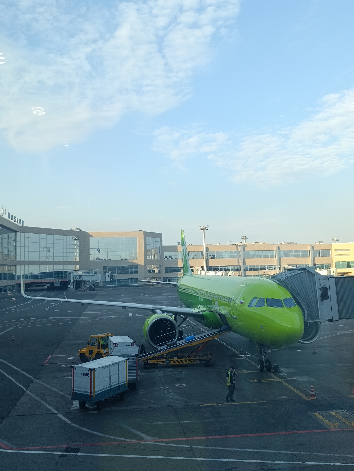 A bright green airplane is parked at an airport gate, connected to a jet bridge. In the background, a modern airport terminal building with large glass windows is visible. Several baggage carts and a small yellow tug vehicle are on the tarmac near the aircraft. A person wearing a high-visibility vest is walking on the tarmac. The scene appears to be at Domodedovo International Airport in Moscow, Russia, given the visible Cyrillic lettering on the building and the distinctive airline livery. The sky is a clear blue with scattered clouds.