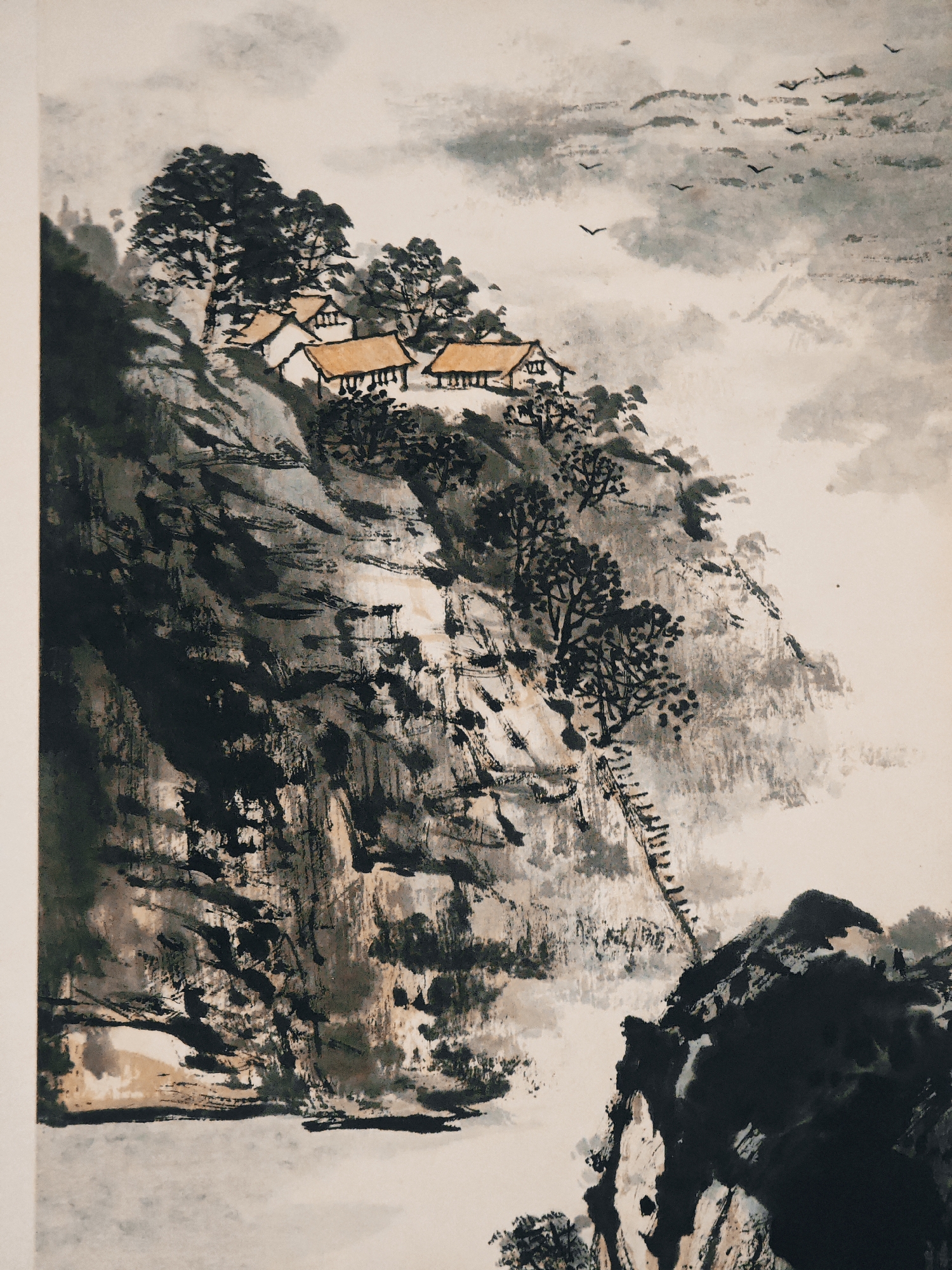This is a traditional Chinese ink wash painting depicting a dramatic and misty mountainous landscape, characteristic of the Jiulongpo District, China. The scene is dominated by a towering, rugged cliff face, rendered with bold, dark ink strokes that convey its steep and imposing nature.
Perched precariously on a ledge within this mountain are several small, white houses with distinctive yellow-orange tiled roofs. These dwellings are surrounded by dense clusters of trees, depicted with dark, intricate brushwork. Further down the cliff, a long, narrow, ladder-like structure ascends a steep section, suggesting a challenging path to the remote settlement.
The background and upper sections of the painting feature soft, diluted ink washes, creating an ethereal mist or cloudy sky that envelops the distant peaks. A flock of dark birds is seen flying across this cloudy expanse, adding a sense of movement and scale to the vast landscape. The overall impression is one of profound tranquility and isolation, emphasizing the majestic and sometimes formidable beauty of nature. The weather appears overcast or misty, contributing to the serene and atmospheric quality of the artwork. No people are visible, and there is no discernible text.