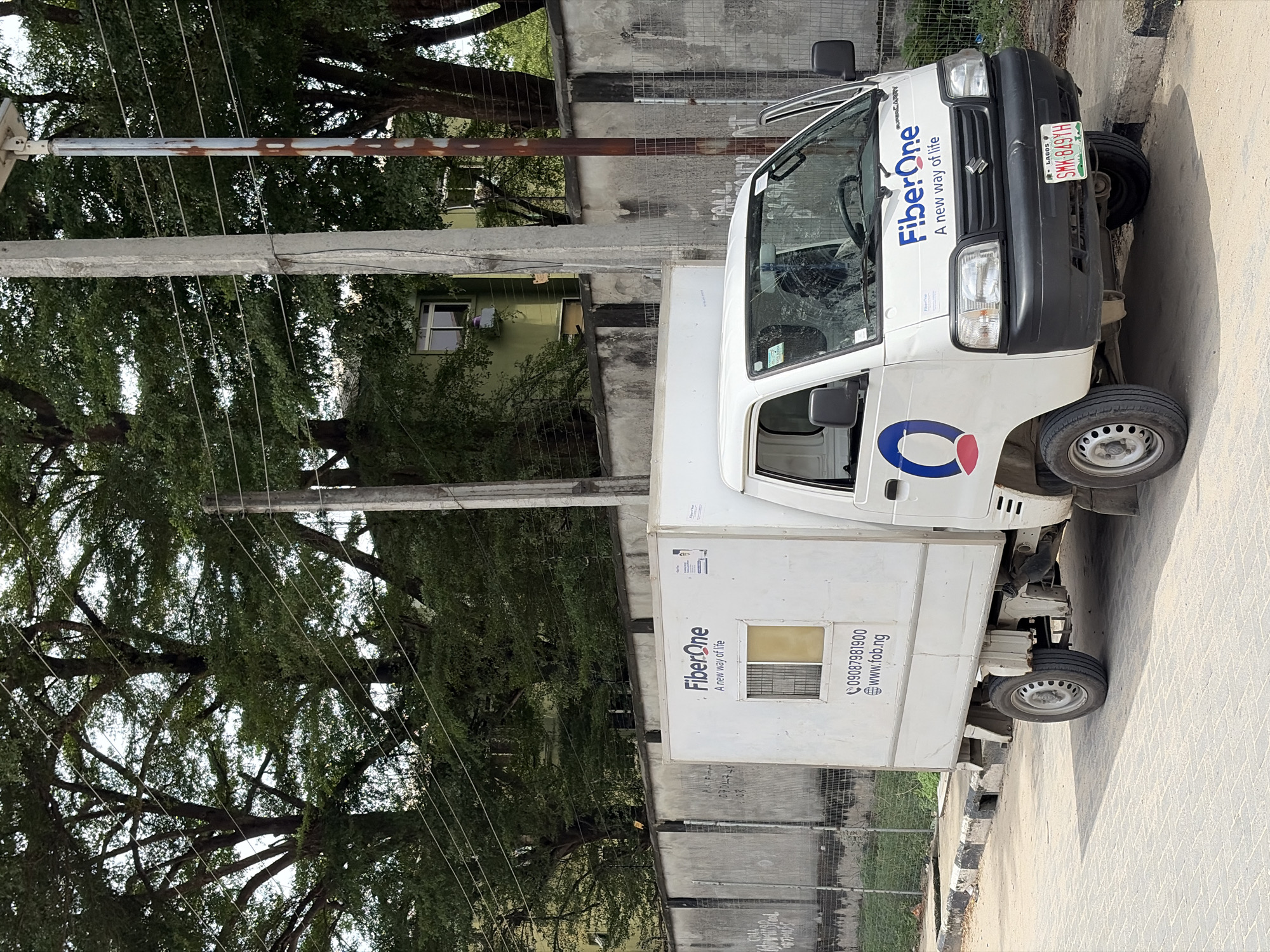 A white utility truck, branded with the "Fiber One" logo and the slogan "A new way of life," is parked outdoors during what appears to be a clear, sunny daytime. The truck is identified as a "SUPERCARRY" model and features a Nigerian license plate that reads "LAGOS SMK-849YH," providing a strong location cue for Lagos, Nigeria, aligning with the Igboefon context. On its side, contact details are visible: "09087981900" and the website "www.fob.ng."
The vehicle is situated on a paved ground surface, adjacent to what appears to be a concrete curb. The background is dominated by large, leafy green trees, suggesting a verdant environment. Behind the trees, a light-colored building with a visible window can be partially discerned. Utility infrastructure, including a concrete pole and a rusty metal pole, runs across the upper part of the scene, supporting multiple overhead wires. The bright lighting indicates good weather, likely late morning or early afternoon. No individuals or specific activities are depicted; the scene focuses on the parked utility vehicle and its immediate surroundings.