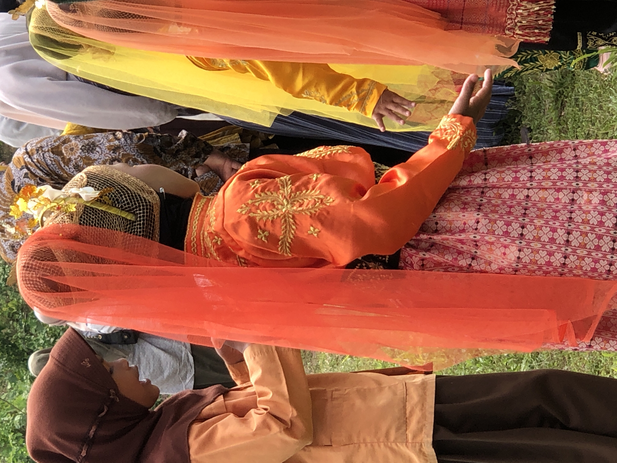 This image, captured in Langkuta, Indonesia, shows a group of people, mostly children, dressed in traditional attire. The focus is on two figures in the foreground. The person on the left wears a brown hijab and an orange tunic with dark brown pants. To their right, a figure in an orange, embroidered outfit with a patterned skirt is partially obscured by a vibrant orange veil. Behind them, other individuals are visible, some in yellow and orange veils, and one wearing a patterned fabric reminiscent of batik. The background consists of lush green foliage. The scene appears to be outdoors, possibly a cultural event or celebration.