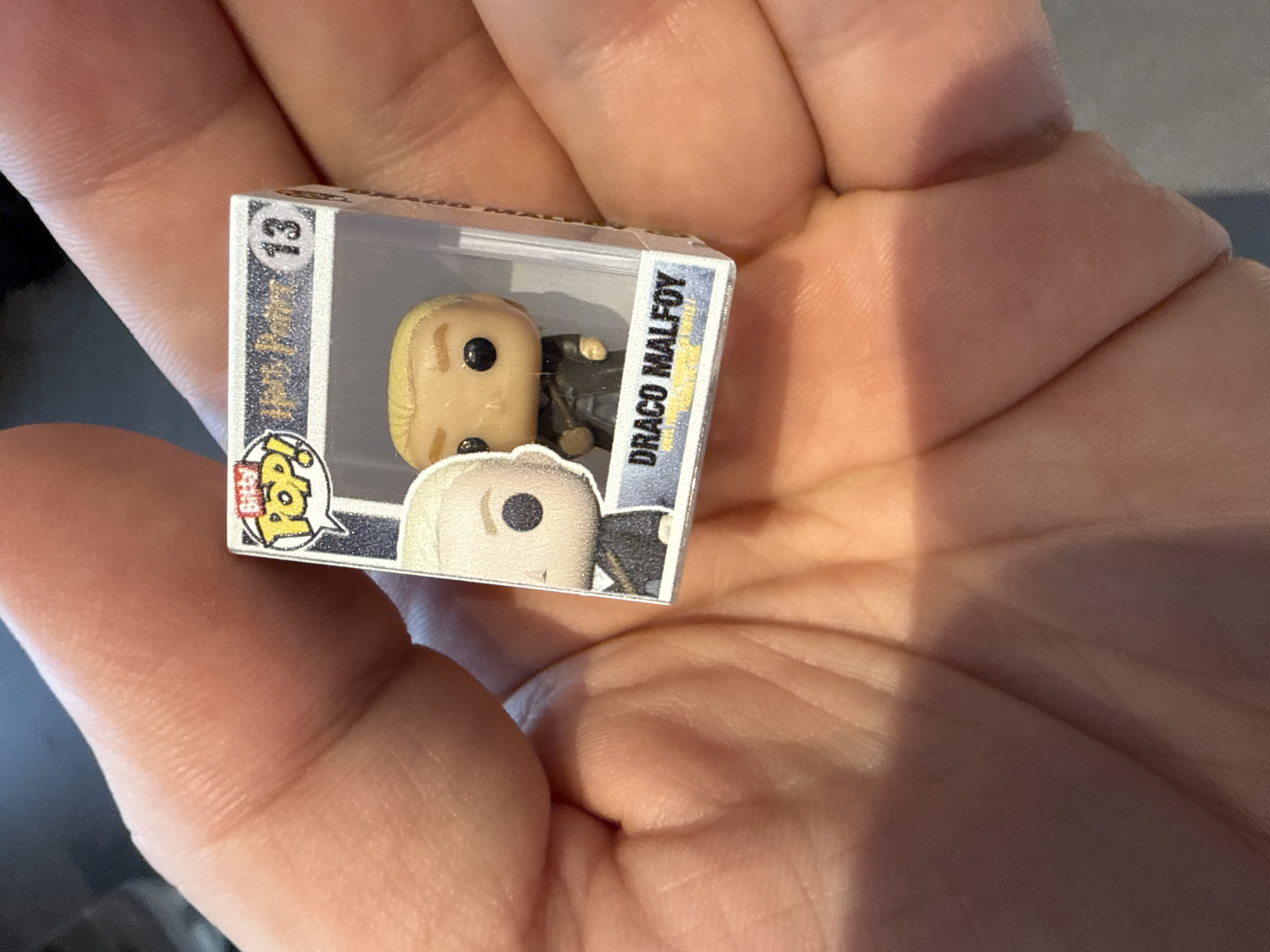 A hand holds a miniature Funko Pop box. The box contains a figure of Draco Malfoy from the Harry Potter series. The box is numbered 13. The environment is undiscernible.