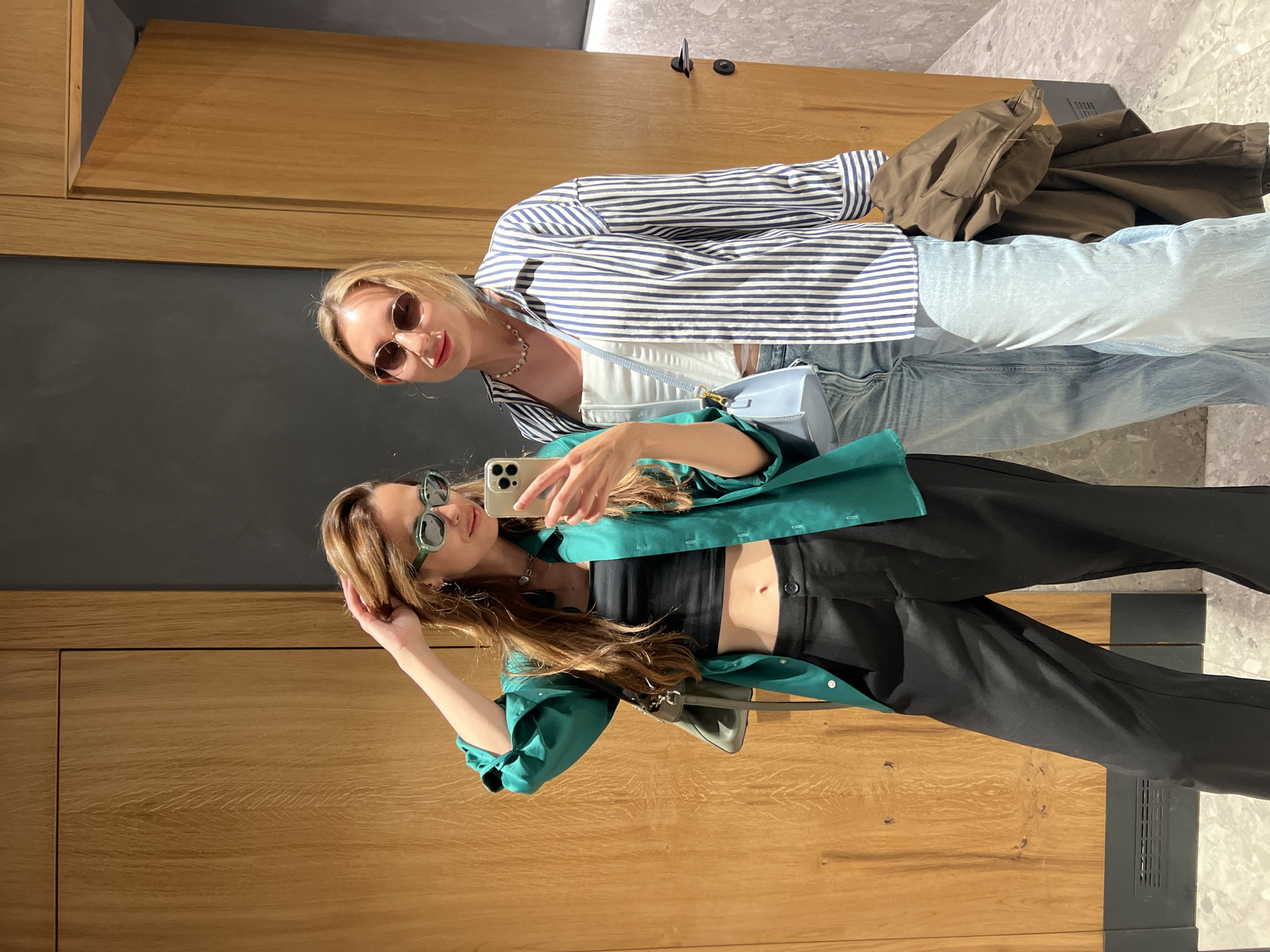 This image captures two young adult females posing for a photograph in an indoor setting, specified as Kuk, Montenegro. The environment presents a modern, minimalist aesthetic with prominent architectural features.
The female on the left has long, light brown hair and wears green rectangular sunglasses. She is dressed in an oversized emerald green button-up shirt, worn open over a black crop top that exposes her midriff and a visible navel piercing. She wears black wide-leg trousers and a silver choker necklace. Her left hand is raised, touching her hair, while her right hand holds a light-colored smartphone with a multi-lens camera array (consistent with recent iPhone models) pointed towards herself, indicating a selfie pose. A light grey shoulder bag is slung over her left shoulder. She exhibits a subtle smile.
The female on the right has blonde hair, pulled back, and wears round, amber-tinted sunglasses. Her attire consists of a blue and white vertically striped oversized button-up shirt, worn open over a white strapless or corset-style top. She wears light blue jeans and a multi-colored beaded necklace. A light blue crossbody bag is visible. She holds a folded brown jacket or coat over her left arm. She displays a slight smile.
The background consists of two flush, light-colored wooden doors with vertical grain, suggesting a modern design. A dark gray wall panel is situated between the doors and extends to the left. The right door is slightly ajar, revealing a darker interior. A dark doorknob and lock are visible on the right door. A black rectangular vent is present at the base of the left wooden door. The floor is covered with large, light gray polished stone or marble tiles featuring subtle veining.