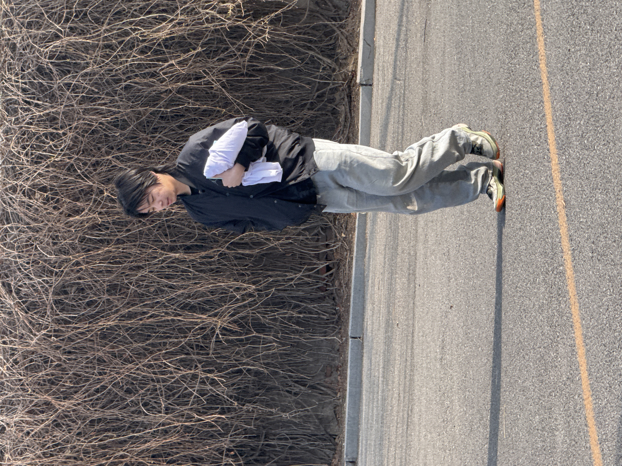 A person stands on a paved surface with a yellow line marking a lane. The person wears gray pants, a black jacket, and multi-colored sneakers and holds a white object in their arms. To the left, a dense thicket of interwoven bare branches extends along the edge of the paved area. The scene's specific location in Zhongtun, China cannot be determined from the image.