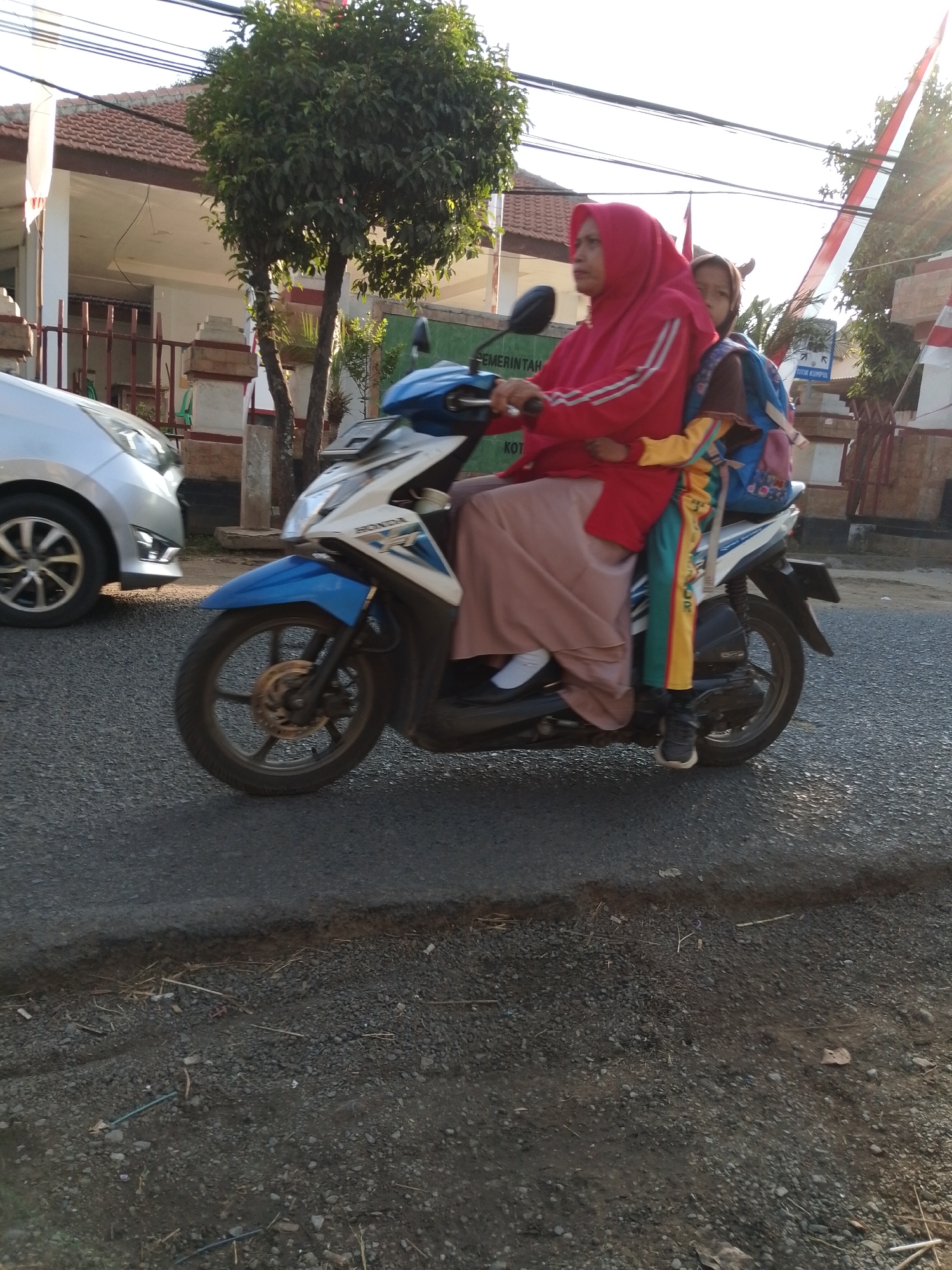 The image shows two individuals, a woman and a child, riding on a blue and white motorcycle on a street in Pasuruan, Indonesia. The woman, wearing a red hijab and jacket, is driving the motorcycle. The child, seated behind her, is wearing a brown hijab and carrying a blue backpack. They are on a paved road, with a silver car visible on the left side of the frame. In the background, there is a building with a tiled roof, a fence, and trees. The scene appears to be set during daytime, with sunlight casting shadows on the road.