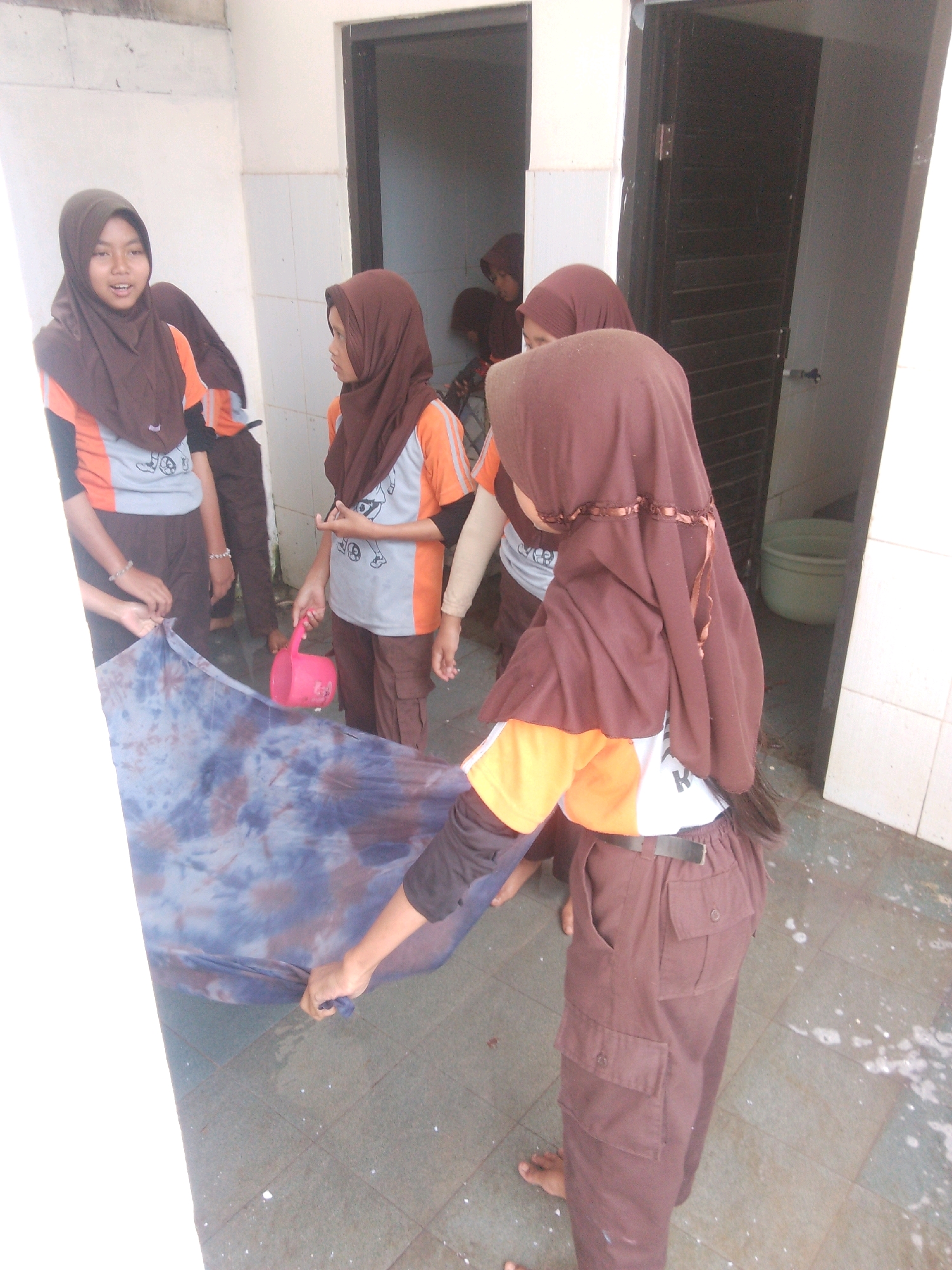 In Betek, Indonesia, a group of young individuals, likely students, are engaged in an activity involving tie-dye fabric. The scene unfolds outdoors, adjacent to a building with white tiled walls and a dark door. One individual, wearing a brown hijab and an orange and gray shirt, holds a pink plastic pitcher and appears to be pouring liquid onto a large piece of tie-dye fabric spread out on the ground. Another individual, also in a brown hijab and a similar uniform, is handling the fabric. The ground is tiled, and there are some wet patches and scattered debris. In the background, partially visible through an open doorway, is another individual wearing a brown hijab, and further back, a green tub is visible. The overall impression is of an informal educational or recreational activity taking place in a communal outdoor space.