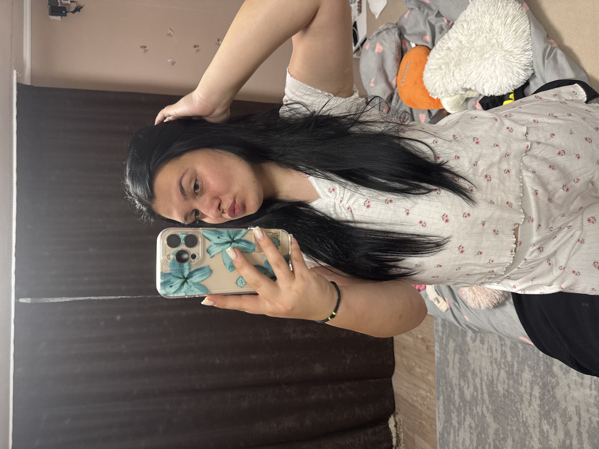 A female individual is captured in a mirror selfie taken indoors in Rogosh, Bulgaria. The subject, a young adult with long, dark hair, is wearing a white two-piece set adorned with small pink floral patterns. Her left hand holds a smartphone with a clear case featuring large blue floral designs, positioned to capture her reflection. Her right hand is raised, with fingers touching the right side of her head. Her lips are pursed in a 'duck face' pose. A black woven bracelet with small yellow beads is visible on her left wrist. The nails on her left hand are manicured with light-colored polish, possibly French tips.
The background visible in the mirror reflection includes dark brown curtains to the left and a light peach-colored wall to the right. Several small, reflective butterfly-shaped decals are affixed to the peach wall. A black electronic device with a blue indicator light is mounted high on the peach wall. The floor features light-colored wood, partially covered by a grey patterned rug. In the lower right foreground, a bed with grey bedding patterned with pink hearts is visible, along with an orange cushion and a white fluffy cushion.