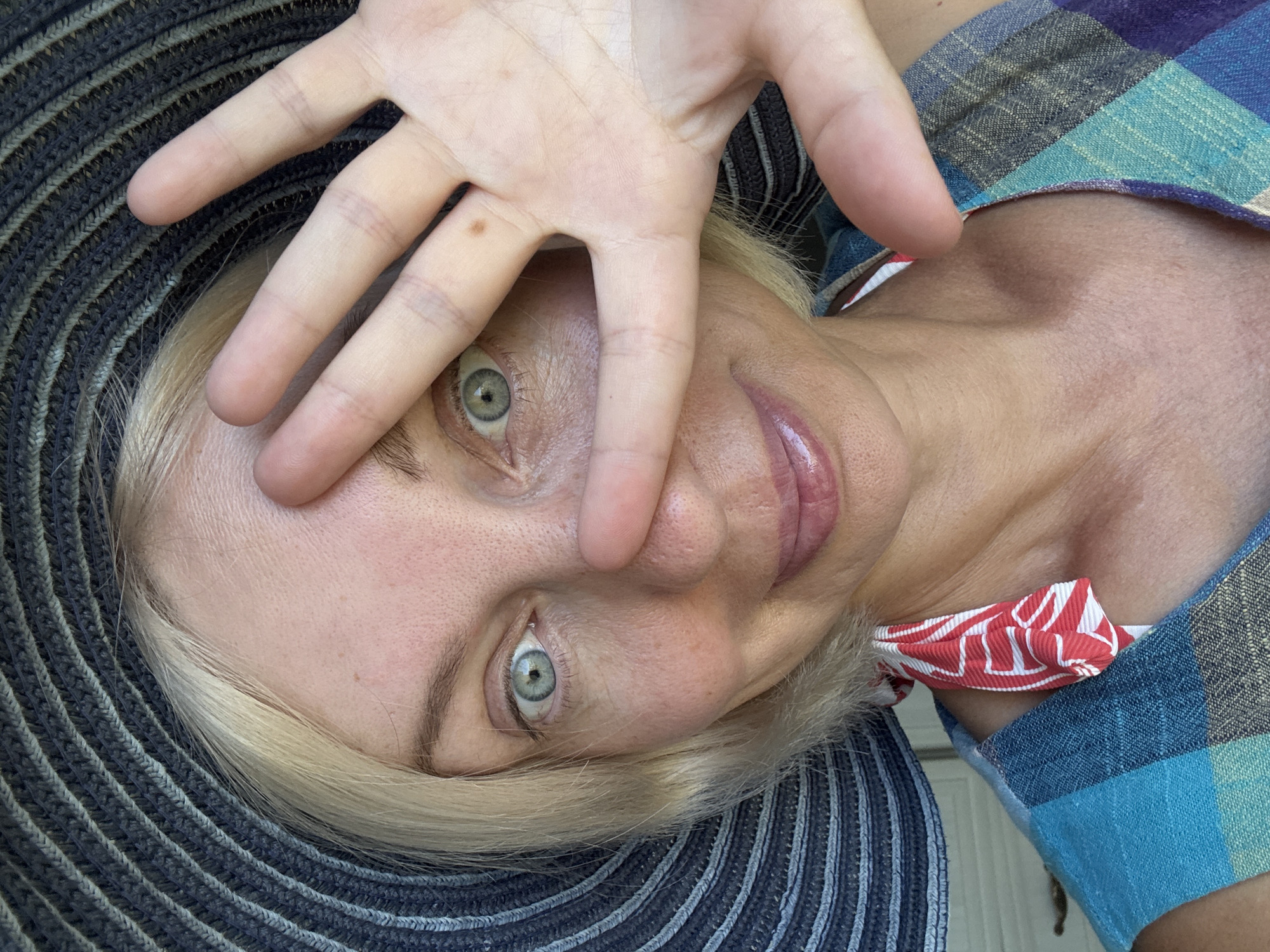 A woman is holding her hand up to her face, partially obscuring her eyes. She is wearing a wide-brimmed hat and a colorful striped dress. The image appears to be a selfie taken outdoors, possibly in a park or garden, with a blurred background suggesting a natural setting. The lighting indicates it is daytime. There is no discernible information linking this image to the city of Sochi, Russia.