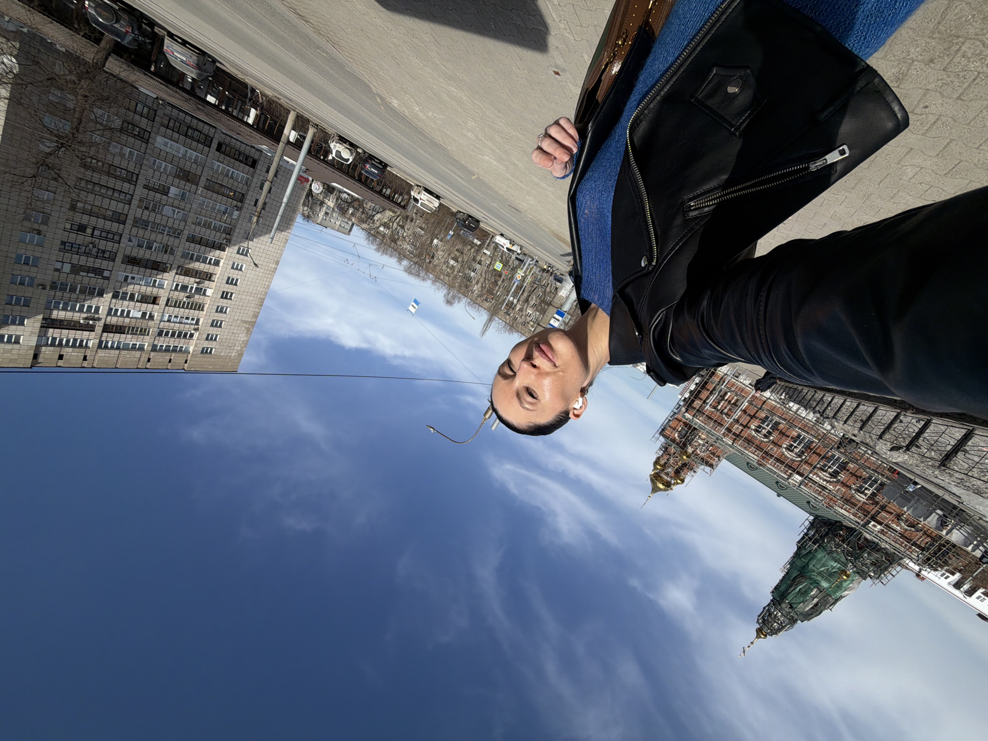 A woman takes a selfie outside on a city street. Buildings are visible in the background. The closest building on the left is gray and has many windows. A second, older building is on the right; its gold spire is under repair. Based on the location of these buildings in Perm, Russia, the building on the right is likely the Perm State Art Gallery housed in the Transfiguration Cathedral, and the grey building on the left is a nearby apartment building.
