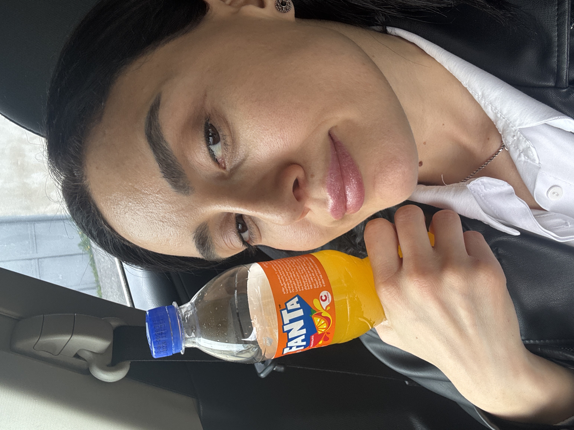 A woman with dark hair is holding a bottle of Fanta orange soda. She is wearing a white shirt underneath a black jacket and a silver necklace. It appears she is in a car. The Fanta bottle label features the brand name in bold letters, along with an orange fruit illustration. The bottle cap is blue. The bottle is partially empty. The image captures the woman from an eye-level perspective. Based on the OCR in the image it is likely that the image was taken in Tbilisi, Georgia.