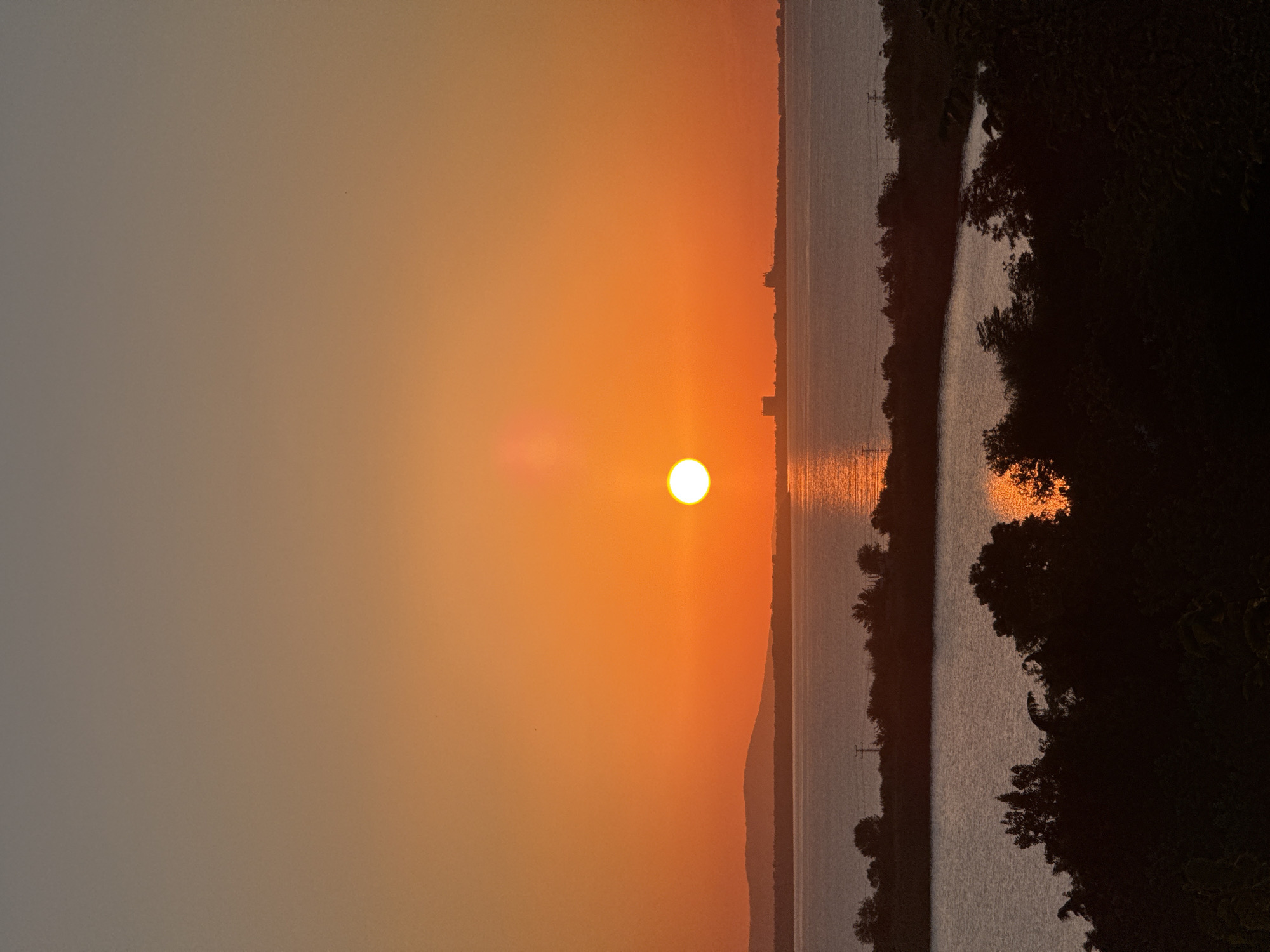 This is a photograph of a sunset over the sea in Burgas, Bulgaria. The sun is a bright, orange orb, low on the horizon, casting a golden reflection on the water. The sky is a gradient of orange and grey. In the distance, a silhouetted coastline is visible, with buildings and hills. The foreground shows dark silhouettes of trees and vegetation. The scene is peaceful and serene, capturing the natural beauty of a sunset.