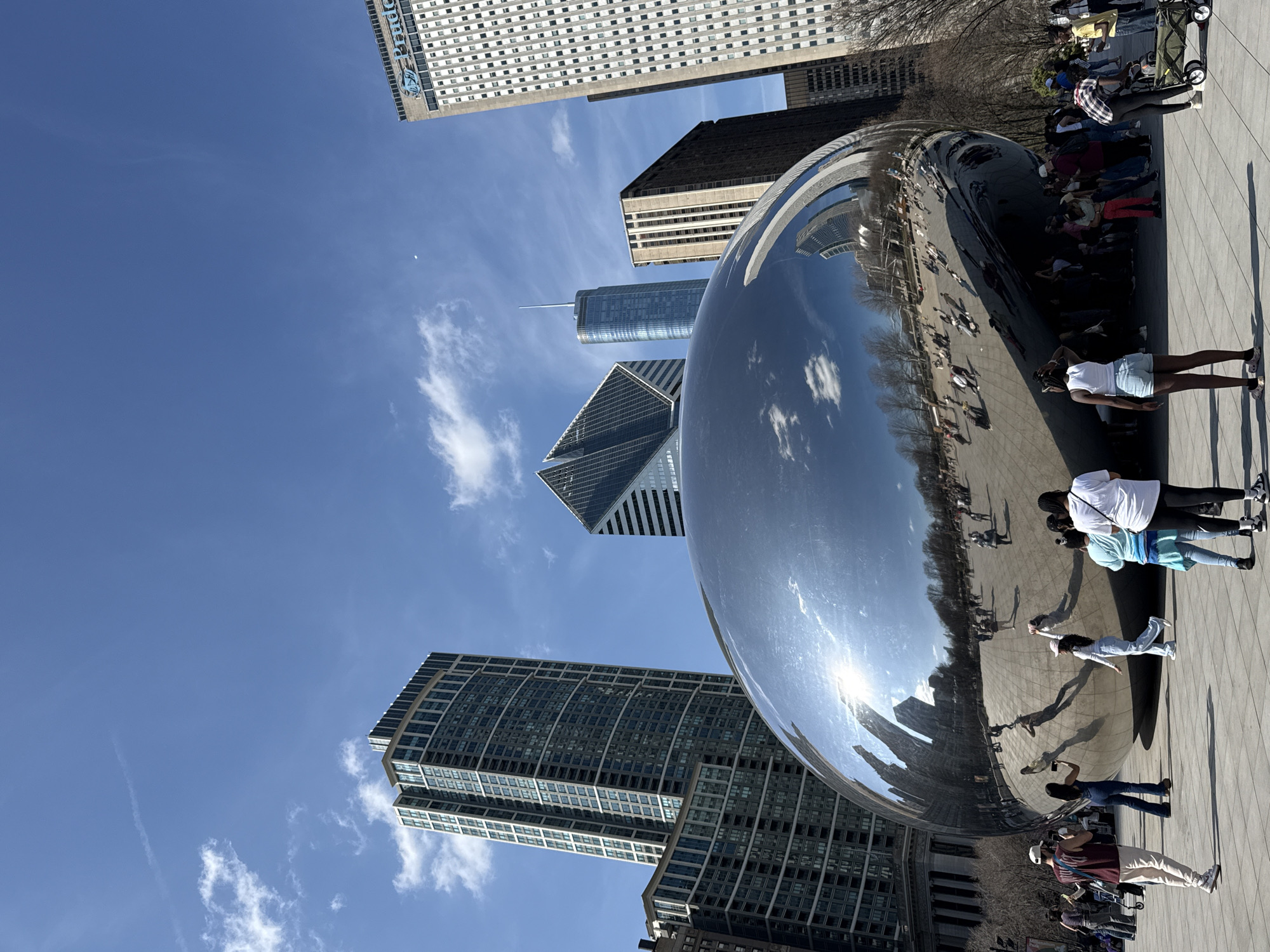 The image depicts a ground-level view of "The Bean," a large, reflective sculpture formally known as Cloud Gate, located in Millennium Park in Chicago, Illinois. The sky is clear and blue with light clouds visible in the background. The sculpture reflects the city's skyline, the surrounding park, and the people visiting. People are interacting with the artwork, taking pictures and walking around it. Several buildings are visible in the background. The photograph is taken from a low angle, emphasizing the sculpture's size and its reflections.