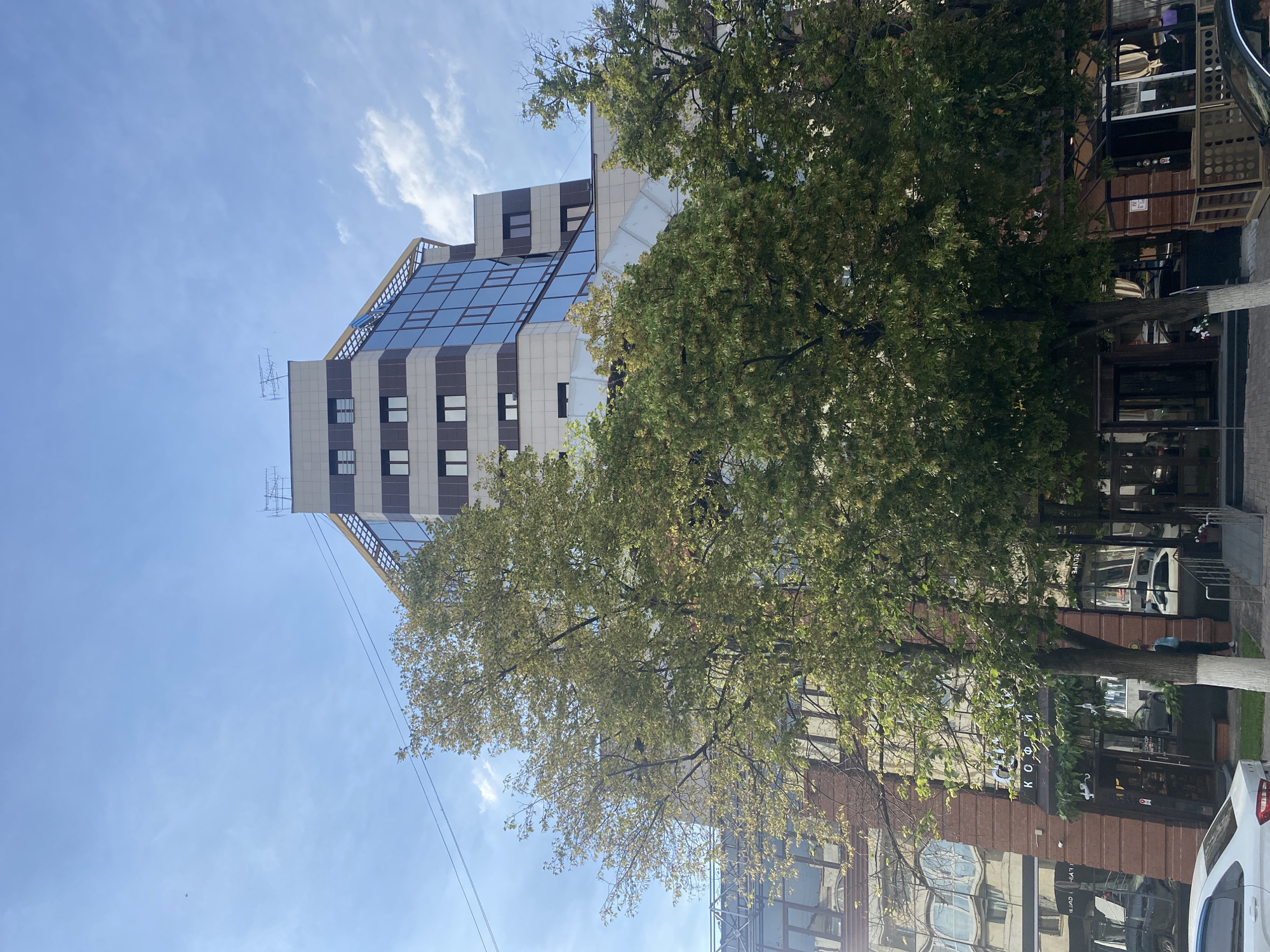 In an urban setting, a contemporary building with a glass facade and horizontal striping stands tall against a partially cloudy blue sky. Mature trees with lush green foliage are positioned in front of the building, partially obscuring its lower levels. The building's upper sections feature a sleek design with multiple windows. In the foreground, a white car is parked on a paved surface. The street level of the building appears to house businesses, with storefronts visible through the glass. A few individuals are seen near the entrance of one of the establishments. The overall scene captures a section of a city on a bright day.