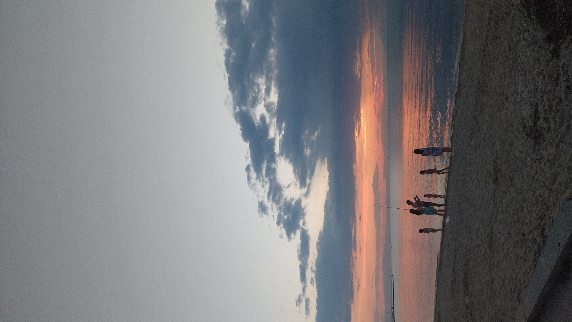 The image depicts a beach scene at sunset in Perea, Greece. A group of six people, consisting of adults and children, are silhouetted against the vibrant sky and calm sea. One adult, possibly a parent, is holding a fishing rod, suggesting a fishing activity. The group appears to be engaged in leisure activities on the sandy shore. The environment is characterized by a beach, the sea, and a colorful sunset with clouds painting the sky in hues of orange, pink, and grey. The overall atmosphere is serene and tranquil, capturing a moment of family time by the sea.
