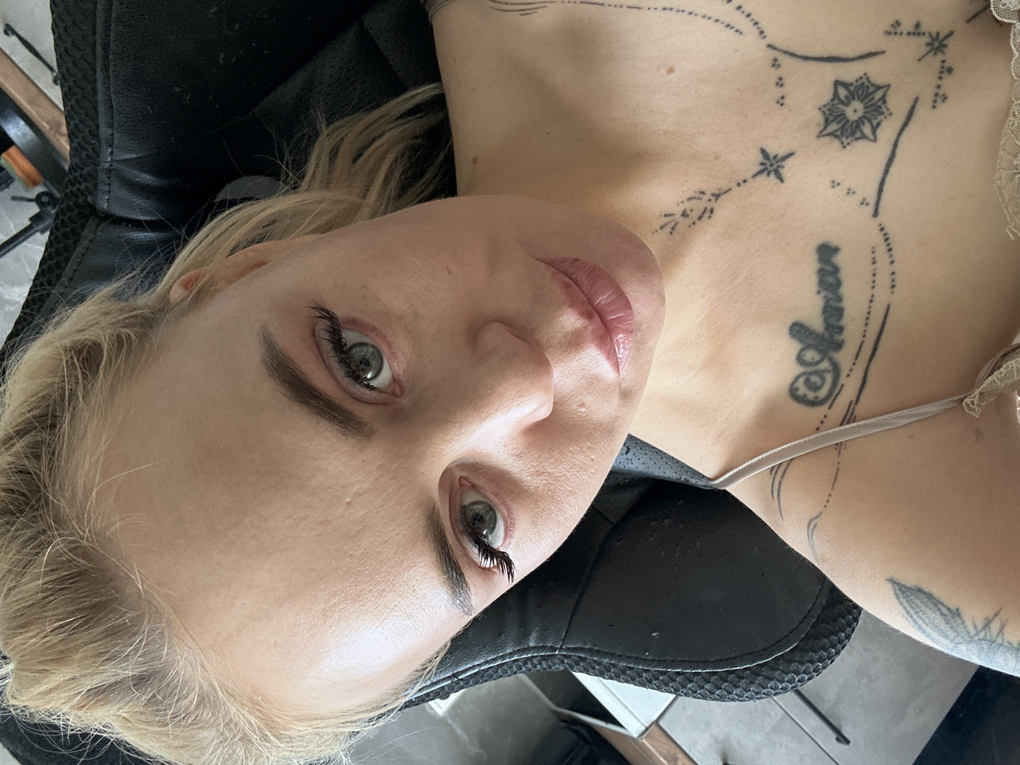 An adult female with fair skin and blonde hair, styled upwards, is captured in a close-up selfie, looking directly into the camera. Her light-colored eyes (appearing blue or grey) are framed by dark eyebrows and prominent false eyelashes visible on her left eye. Her lips are slightly parted, coated in a light pink gloss or lipstick, with a neutral to slight smile. Numerous tattoos are visible on her upper torso: a script tattoo, possibly reading "Aman" or "Annien," with decorative swirls and dots across her left clavicle area, and on her right upper chest, a design featuring stars and a multi-petaled, mandala-like flower connected by dotted lines. A faint, possibly winged or feathered tattoo is visible on her left upper arm/shoulder. She is wearing a light-colored, thin-strapped top, possibly with lace detailing visible at the bottom edge of the frame. The woman is seated and reclined against a black upholstered chair, possibly a gaming or office chair, with a textured backrest and headrest visible. Indistinct background elements suggest an indoor setting, including dark objects at the upper right and light-colored floor tiles and a dark object at the lower left. The location is an indoor room in the city of Unknown, Unknown.