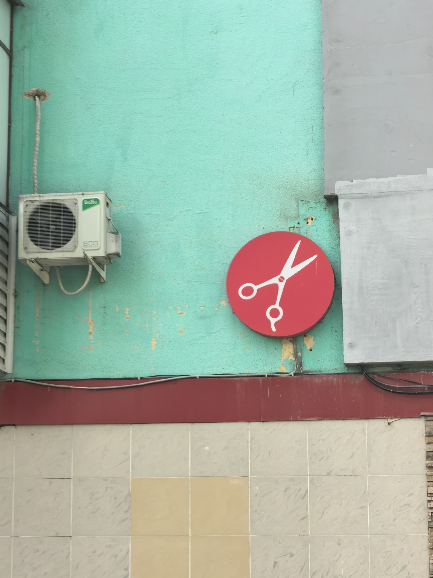 A vertical shot shows the exterior of a building. An air conditioning unit is attached to the upper left green wall. To the right is a circular red sign with a white scissor symbol, indicating a barbershop. Below, a horizontal red band is visible, topped by beige tiles. This is in Sterlitamak, Russia.