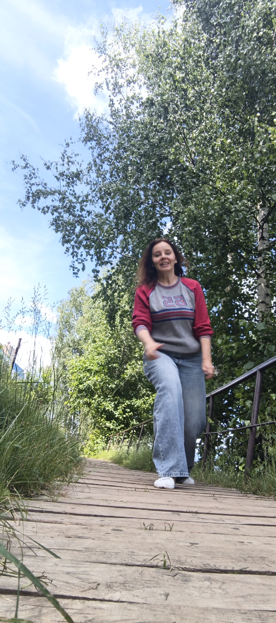 A young woman with medium-length brown hair smiles at the camera as she stands on a wooden walkway. She is wearing a grey sweatshirt with red sleeves and a blue and red striped design across the chest, and loose-fitting blue jeans. Her feet are clad in white sneakers. The walkway appears to be part of an outdoor stairway or path, with a metal handrail visible to her right. The background is dominated by lush green trees and a bright blue sky with scattered clouds, suggesting a sunny day. The overall scene evokes a sense of cheerful outdoor activity. The text on her sweatshirt appears to be the number "23" in a stylized font. The location is Chaikovsky, Russia.