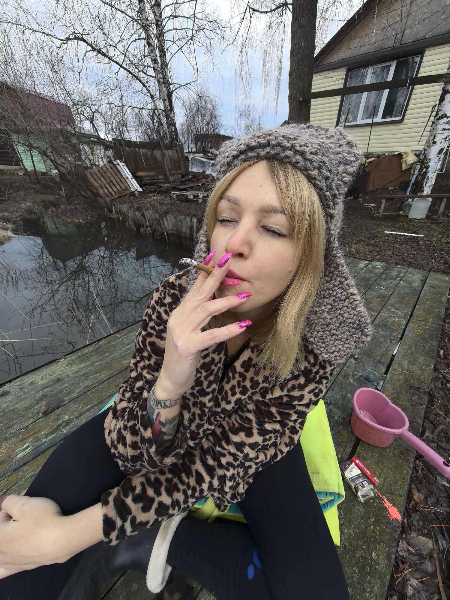 A woman with blonde hair, fair skin, and closed eyes is pictured from a high-angle perspective, smoking a cigarette outdoors in Noviy, Russia. She is seated on a weathered wooden plank dock or platform. Her long nails are painted a vibrant, glittery pink, matching her lipstick. She wears a thick, chunky knit grey hat (possibly with earflaps), a brown and black leopard print fleece jacket, and black leggings, with a bright neon yellow/green garment visible underneath. Tattoos are partially visible on her left forearm. A lit cigarette is held between her pink-manicured fingers.
On the wooden surface next to her are a pink heart-shaped plastic scoop, an open pack of "WINSTON" cigarettes displaying a Russian health warning, and a red lighter.
The scene depicts a rural or semi-rural landscape. A still body of water, possibly a pond or flooded area, reflects the bare branches of trees and an overcast sky. In the background, several rustic wooden houses are visible, including one with light-colored siding and a white window, and another with a dark roof and green wall. Bare birch trees and other deciduous trees, indicative of late winter or early spring, dominate the distant landscape. Wooden fences, debris, and muddy ground contribute to a rustic environment. The time of day is daytime, under a cloudy sky. The woman's closed eyes convey a moment of contemplation or relaxation.