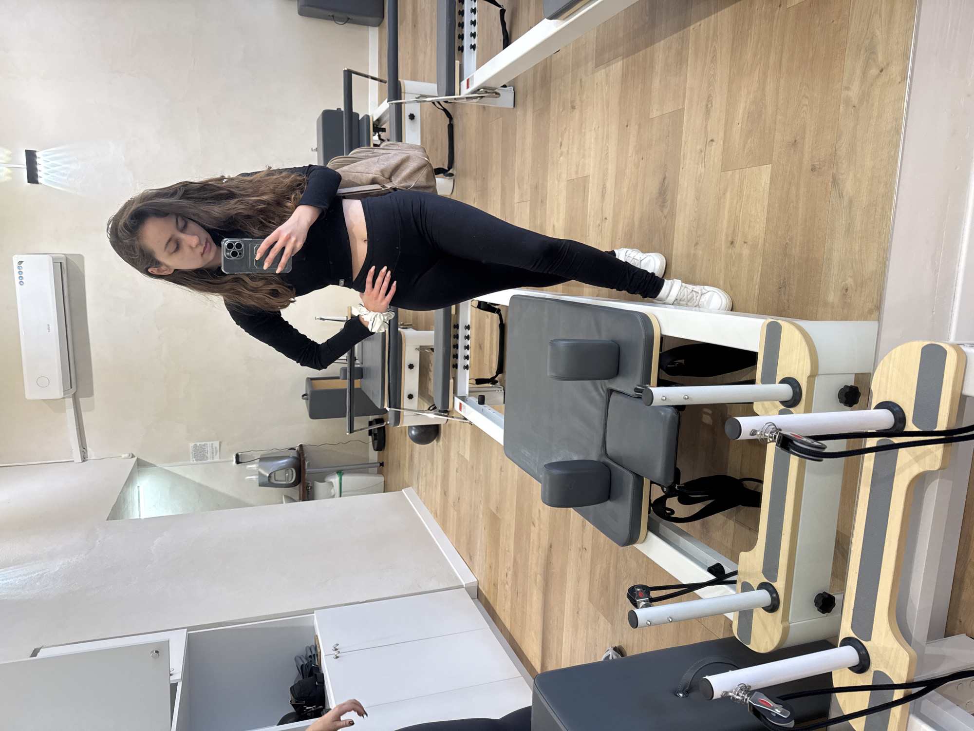 A woman in black athletic wear stands in a Pilates studio in Ramat Gan, Israel. She faces a mirror, using her phone to take a picture. The studio has wooden floors, white walls, and Pilates equipment including reformers and a trapeze table. The woman has long, wavy brown hair.