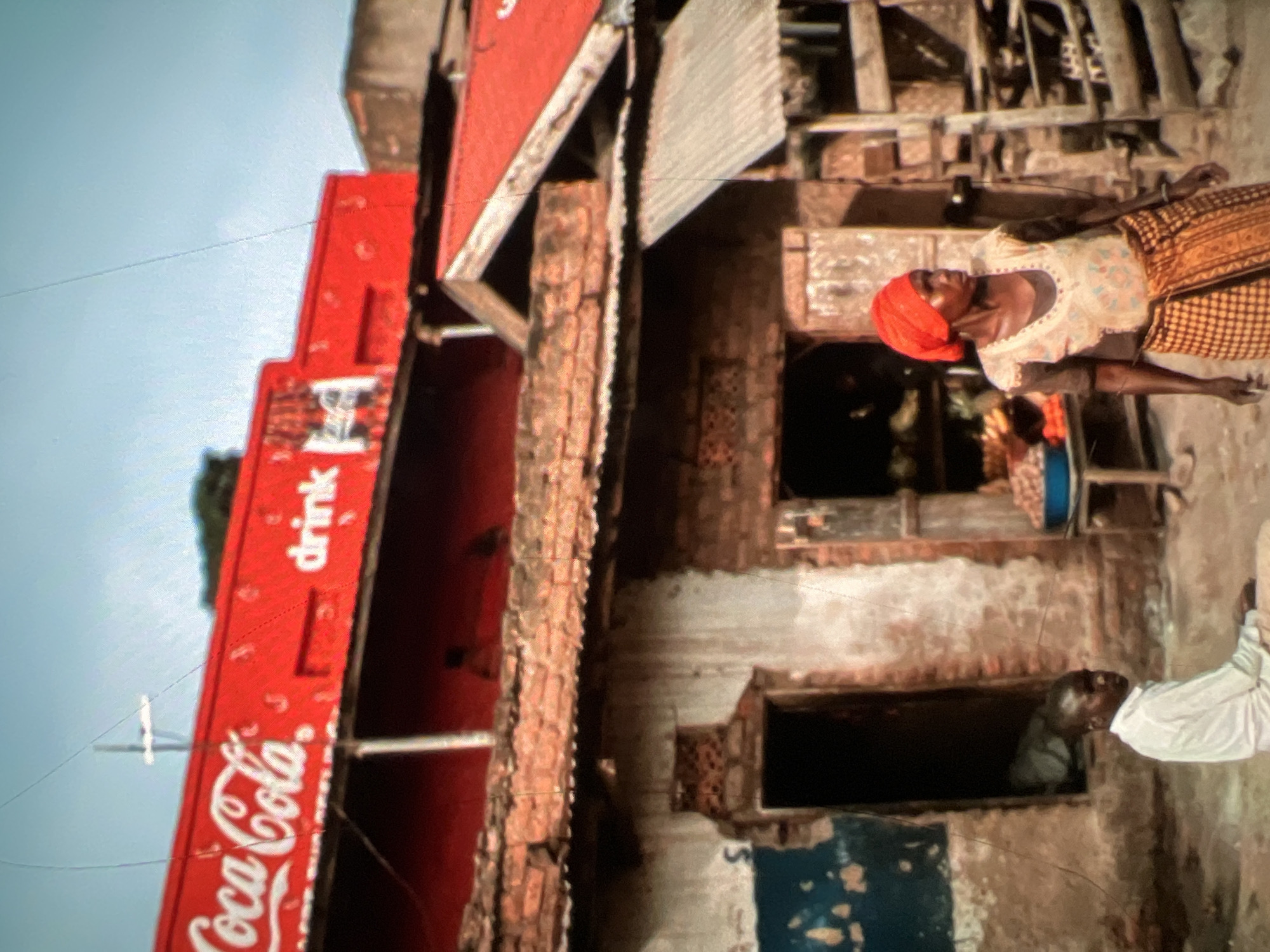 In Lagos, Nigeria, a Coca-Cola branded building with a red facade and white lettering dominates the upper portion of the image, featuring the iconic "Coca-Cola" logo and the word "drink." Below this, a weathered brick building with a corrugated metal roof shows signs of age and disrepair, with a dark, open doorway and a rough plastered exterior. A young boy in a white shirt sits on the ground in front of the doorway. To the right, a woman wearing a red headscarf and a patterned skirt stands in front of a stall selling goods. The scene depicts a bustling street environment with a blend of commercial advertising and everyday life in what appears to be a vibrant urban setting.