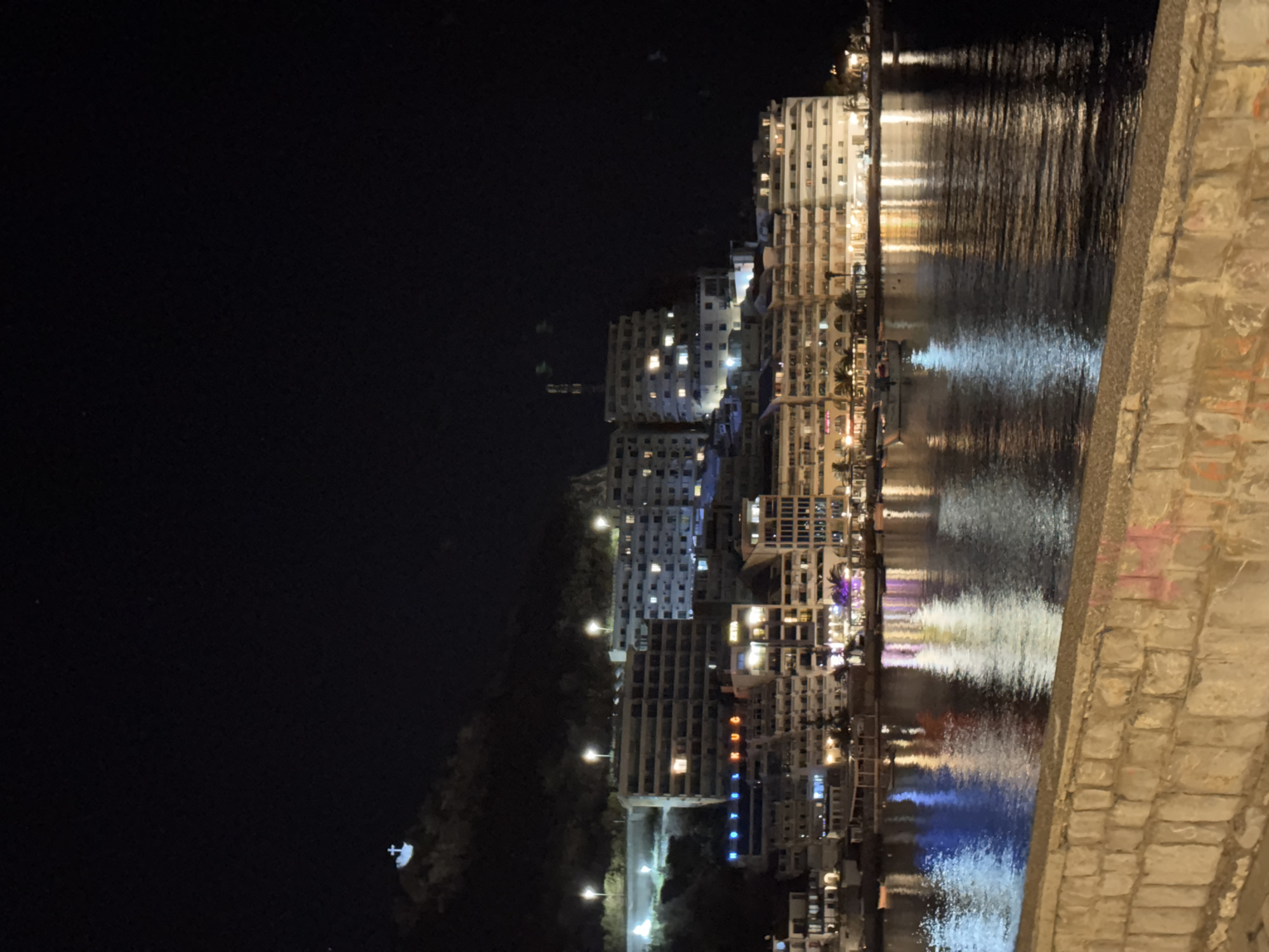 The image shows a nighttime view of Rafailovići, Montenegro, from a slightly elevated perspective, possibly a bridge or embankment. The scene is dominated by the illuminated buildings of the town, reflected in the water. The buildings range in height, with some taller structures visible towards the center of the frame. The lights from the buildings and street lamps create shimmering patterns on the water's surface. The left side of the image is dark, with some vegetation and hints of a hill or mountain. The right side of the image includes a stone wall or structure, indicating the viewpoint. There are no people visible.