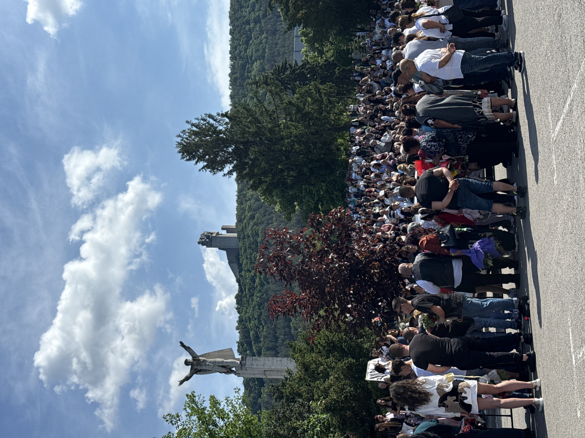 A crowd of people stands on an asphalt surface, likely a road or a square, facing the Monument to 1300 Years of Bulgaria in Shumen, Bulgaria. The monument, featuring a large sculpture of a figure with outstretched arms, is visible in the background against a backdrop of a blue sky with scattered clouds. The trees and the mountain are also visible in the background. The crowd appears to be paying respects at the monument.