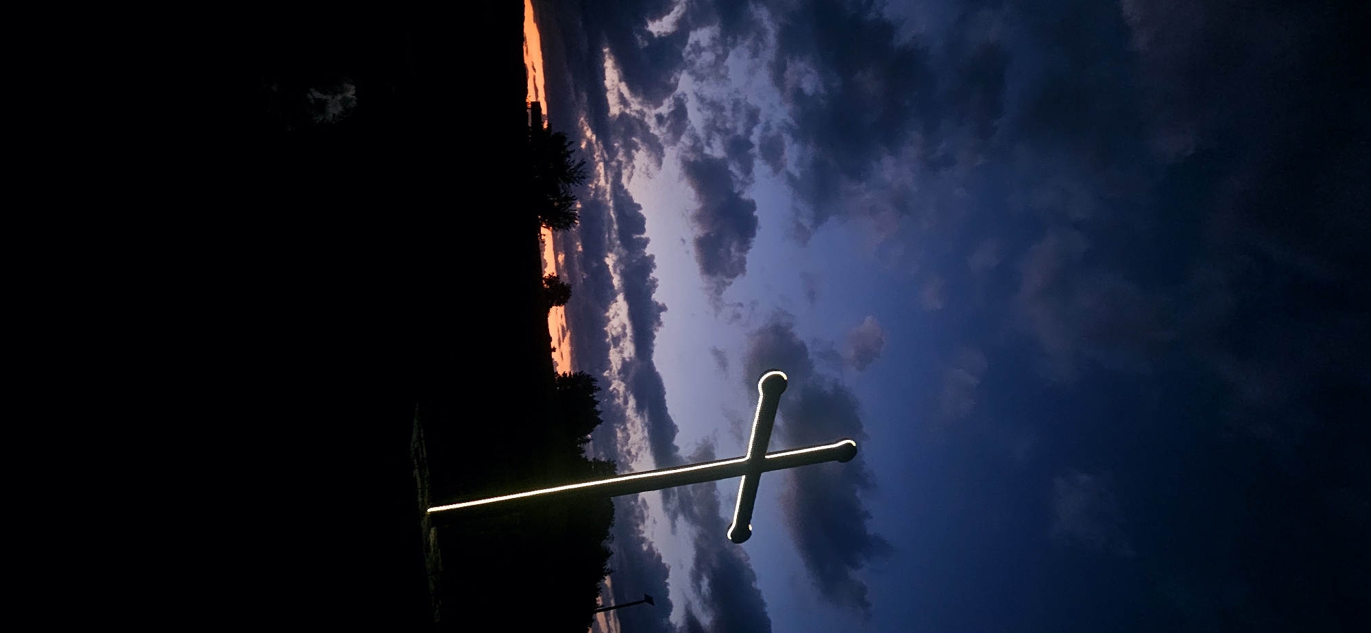 The image depicts an outdoor twilight scene in Gutsal, Bulgaria, featuring a large, illuminated cross. The upper two-thirds of the frame are dominated by a dark blue and indigo sky, characterized by numerous heavy, grey clouds. A band of orange and reddish light from the setting sun is visible along the distant horizon, partially obscured by the cloud cover.
A prominent, tall Christian cross, emitting a bright white light from its perimeter, stands angled slightly left in the right half of the image. The light sharply outlines the cross against the darker sky and foreground. The lower portion of the image, approximately the bottom half, is almost entirely silhouetted in deep black, representing the ground, distant rolling hills, and a dense line of trees on the horizon. No specific details of the landscape are discernible due to the darkness. The base of the illuminated cross is rooted within this dark foreground. No people are present. A faint, unlit pole or structure is barely visible on the far right horizon, within the treeline silhouette. No actions or interactions are depicted.