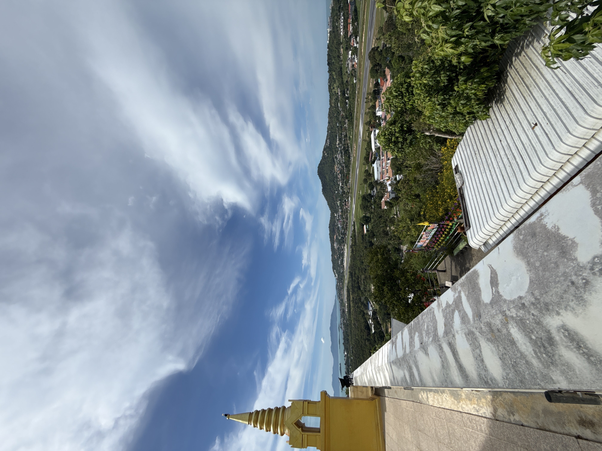 The image provides an elevated view of a landscape in Ban Bang Rak, Thailand. In the foreground on the left, a golden Buddhist stupa is partially visible, perched on a concrete structure. A weathered, light-colored concrete wall extends from the foreground to the middle ground. Beyond the wall, the scene unfolds to reveal a lush green expanse of trees and vegetation. In the middle distance, a runway is clearly visible, indicative of an airport. Scattered houses and buildings are interspersed with the greenery, suggesting a populated area. Further in the background, rolling hills rise, with some topped by trees. In the far distance, a body of water, likely the sea, is visible, with islands or larger landmasses on the horizon. The sky is a vibrant blue, punctuated by a scattering of white, wispy clouds.