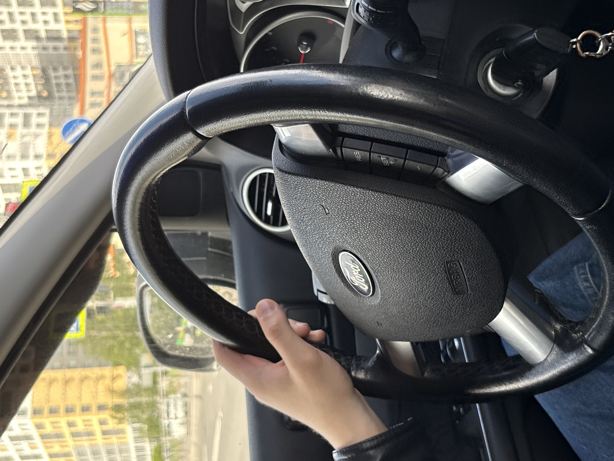 A driver is operating a Ford vehicle in Saint Petersburg, Russia. The driver's left hand is visible on the steering wheel, and a view through the windshield shows buildings and streets of the city. The car's keys are visible in the ignition.