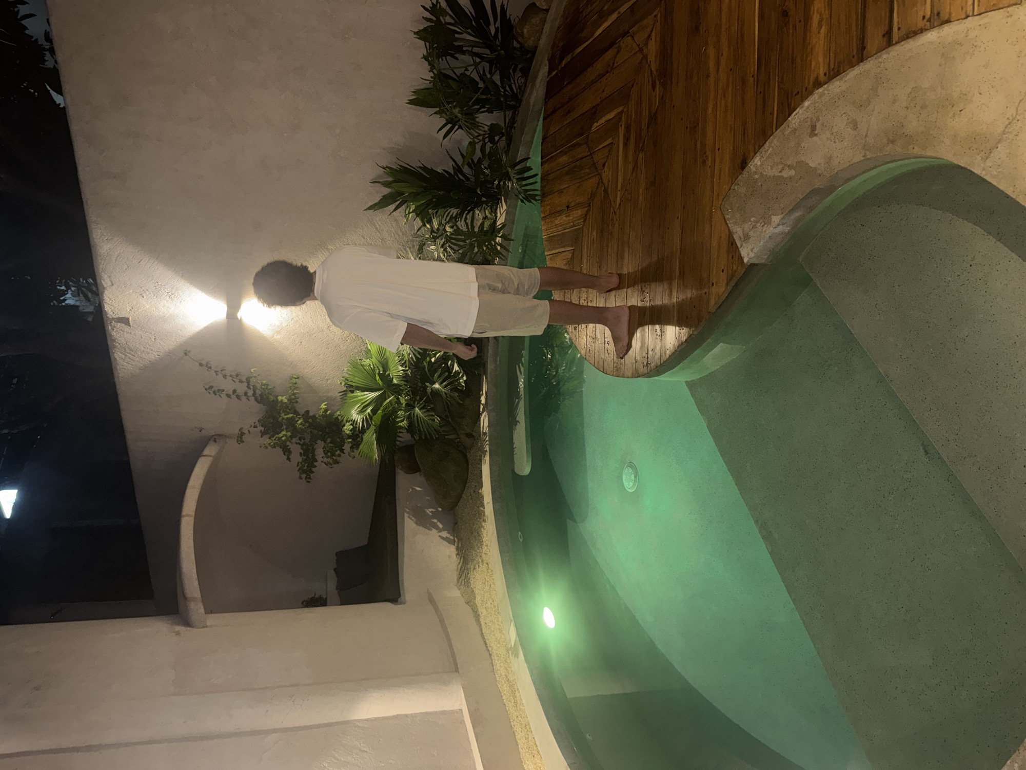 An outdoor scene at night in El Nido, Philippines, depicts a private swimming pool area enclosed by light-colored, textured walls. A lone individual, viewed from behind, stands barefoot on a curved wooden deck bordering a segment of the pool. The person wears a white short-sleeved t-shirt and light-colored shorts, possessing dark, curly hair. Their posture indicates they are observing the illuminated wall and surrounding tropical foliage.
The pool features light blue-green water, brightly lit by submerged green lights, with concrete steps descending into it on the left. A circular drain is visible on the pool floor. The wooden deck, characterized by radial planking, is visibly wet. Adjacent to the pool and extending to the wall, a bed of light-colored gravel separates the water from various green plants, including large-leafed tropical specimens and smaller shrubs, interspersed with large, dark rocks.
A prominent arched alcove is present in the wall on the left side of the scene, appearing dark within. A wall-mounted light fixture, emitting light upwards and downwards, illuminates the wall directly behind the standing individual. The overall ambiance is dimly lit, with artificial light sources providing focal illumination.