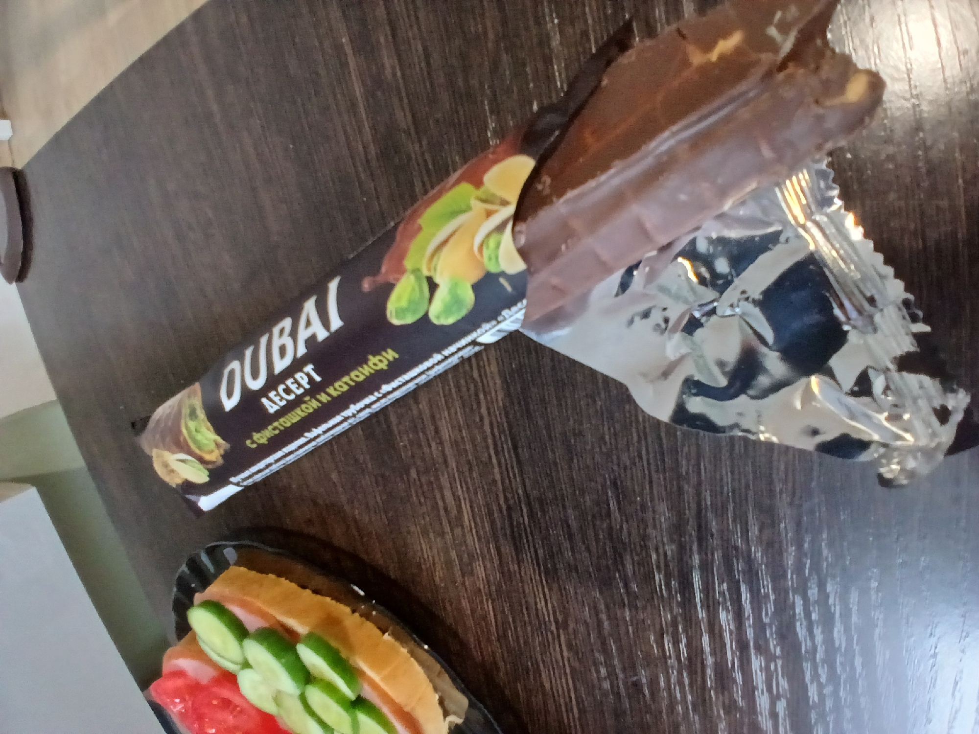 In Yekaterinburg, Russia, a close-up shot captures a wooden table surface where a half-eaten chocolate bar wrapper and the chocolate bar itself are placed. The chocolate bar, partially unwrapped, reveals its glossy, dark brown surface. The wrapper, a metallic foil with printed text and an image of pistachios, lies beside it. To the left of the chocolate bar, a small sandwich is visible on a black plate, topped with slices of tomato and cucumber on bread. The background shows a blurred hint of a room interior, suggesting an indoor setting.
