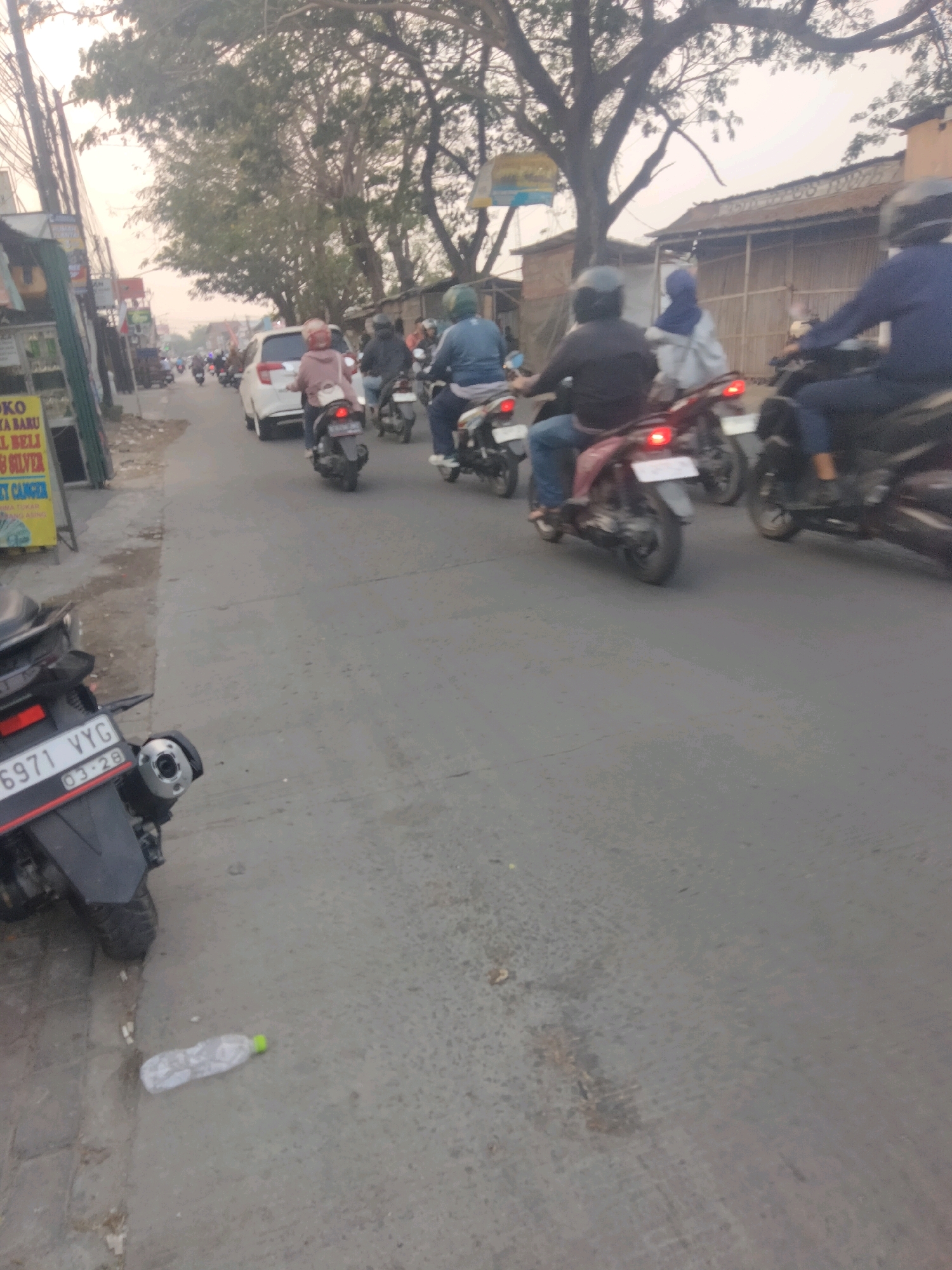 The image shows a street scene in Sepatan, Indonesia, with several motorbikes and a car. In the foreground on the left, the rear of a black motorbike is visible, displaying a license plate that reads "6971 YYG 03.28". To the left of the motorbike is a sidewalk with some debris, and a discarded plastic bottle with a green cap lies on the road. The street is paved and appears to be in moderate condition.
Several motorbikes are moving down the street. On one motorbike, a person wearing a pink headscarf and a light-colored jacket is riding. Another motorbike has a rider in a blue jacket and helmet. Further down the road, a white car is visible. On other motorbikes, riders are wearing helmets and casual clothing, with some appearing to be carrying passengers.
The environment suggests a commercial or residential area. Buildings line the right side of the street, with some appearing to be shops or small businesses, indicated by signage. Trees with sparse foliage are present along the sides of the street and in the background. The sky is overcast, and the overall lighting suggests it might be late afternoon or early morning. The scene depicts typical daily traffic and street life in Indonesia.