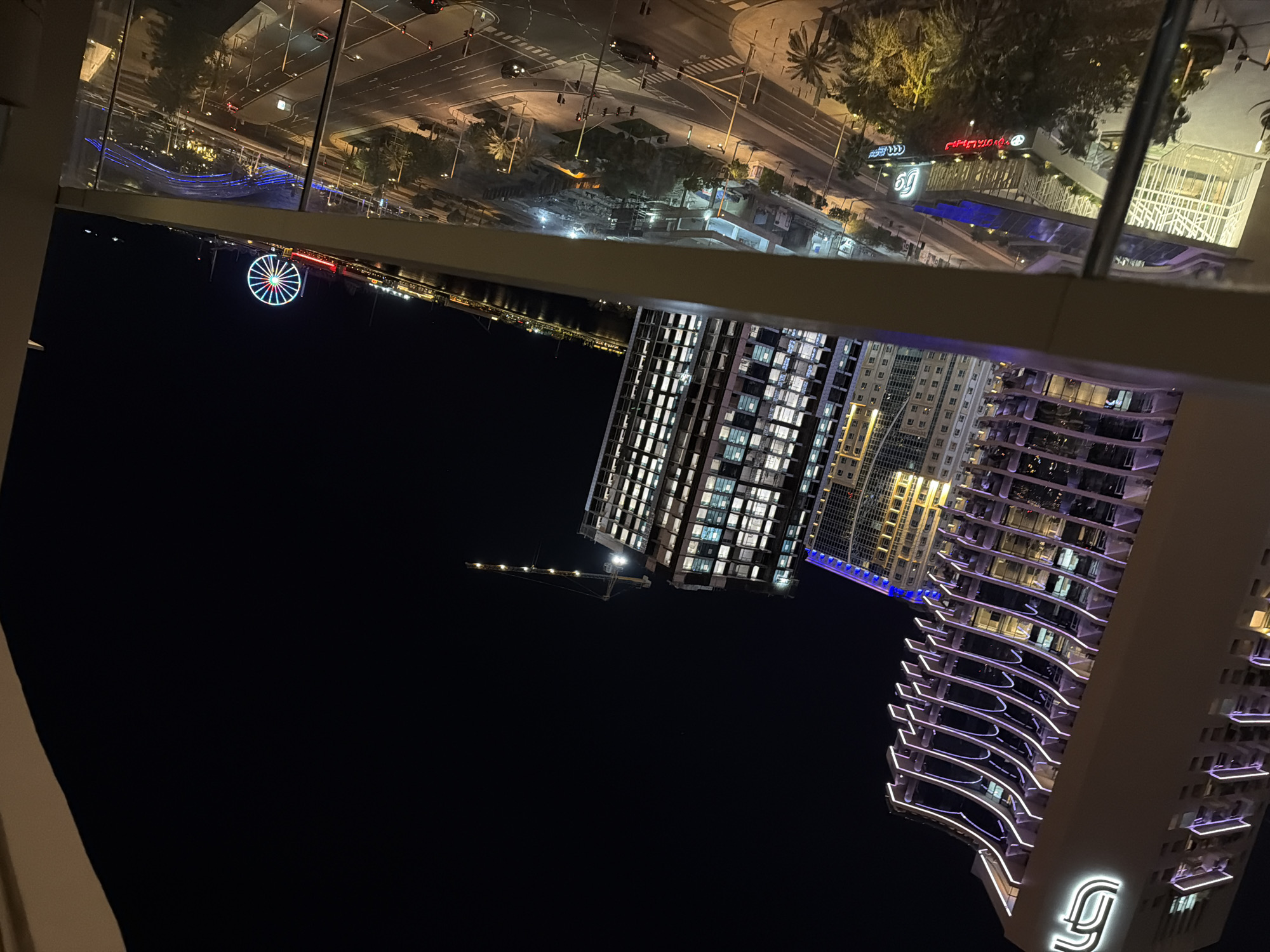 The image presents a detailed night-time panoramic view of a vibrant urban landscape, captured from an elevated perspective, likely a building balcony. The location is Jumeirah Beach Residence (JBR) in Dubai, United Arab Emirates.
In the immediate foreground, a balcony railing and glass barrier are visible, indicating the viewpoint. To the left, a prominent, tall skyscraper features a distinctive, stylized "FJ" or "JG" logo at its top, with its curved balconies elegantly outlined by glowing purple neon lights. Adjacent to it, other modern high-rise buildings are illuminated, showcasing their architectural details.
Below the observer, a bustling street scene unfolds, characterized by a complex intersection of multi-lane roads. Numerous vehicles are in motion, their lights creating streaks on the asphalt. The streets are well-lit by streetlights and bordered by sidewalks and numerous palm trees. At ground level, various commercial establishments are visible, with illuminated signs, some featuring Arabic script, including recognizable names such as "JBR" and "The Walk". A modern building with a distinctive glass-roofed structure is also present at street level.
In the mid-ground and background, a large body of water, likely the Arabian Gulf, separates the observer from the iconic Ain Dubai Ferris wheel, which stands brightly illuminated with a spectrum of colors. Lights from other structures and the distant cityscape reflect on the water's surface. A tall construction crane is also visible against the dark night sky, hinting at ongoing development in the area. The scene is devoid of clearly discernible individual people, focusing instead on the city's architectural and infrastructural elements.