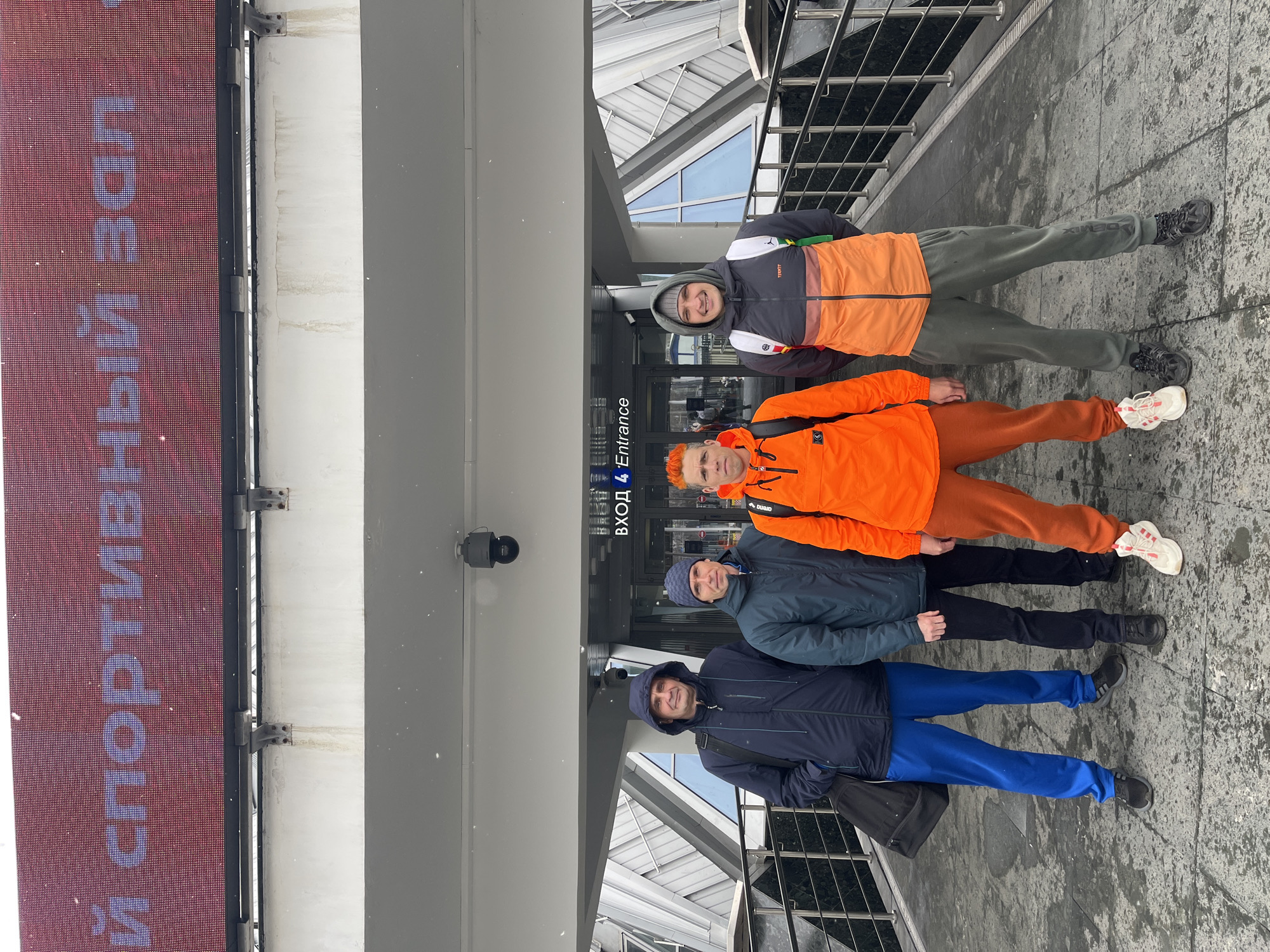 Four men stand together near an entrance marked "ВХОД * Entrance," likely at a sports complex in Moscow, Russia, as suggested by the sign reading "Спортивный Зал" (Sports Hall) in Russian. The men are dressed in casual, cold-weather clothing, including jackets and tracksuits. The setting appears to be outdoors, possibly on a paved walkway leading to the entrance of the building. The architectural design of the building suggests a modern style. The sky is overcast, and light appears diffused, indicating possibly wintry or cloudy conditions.