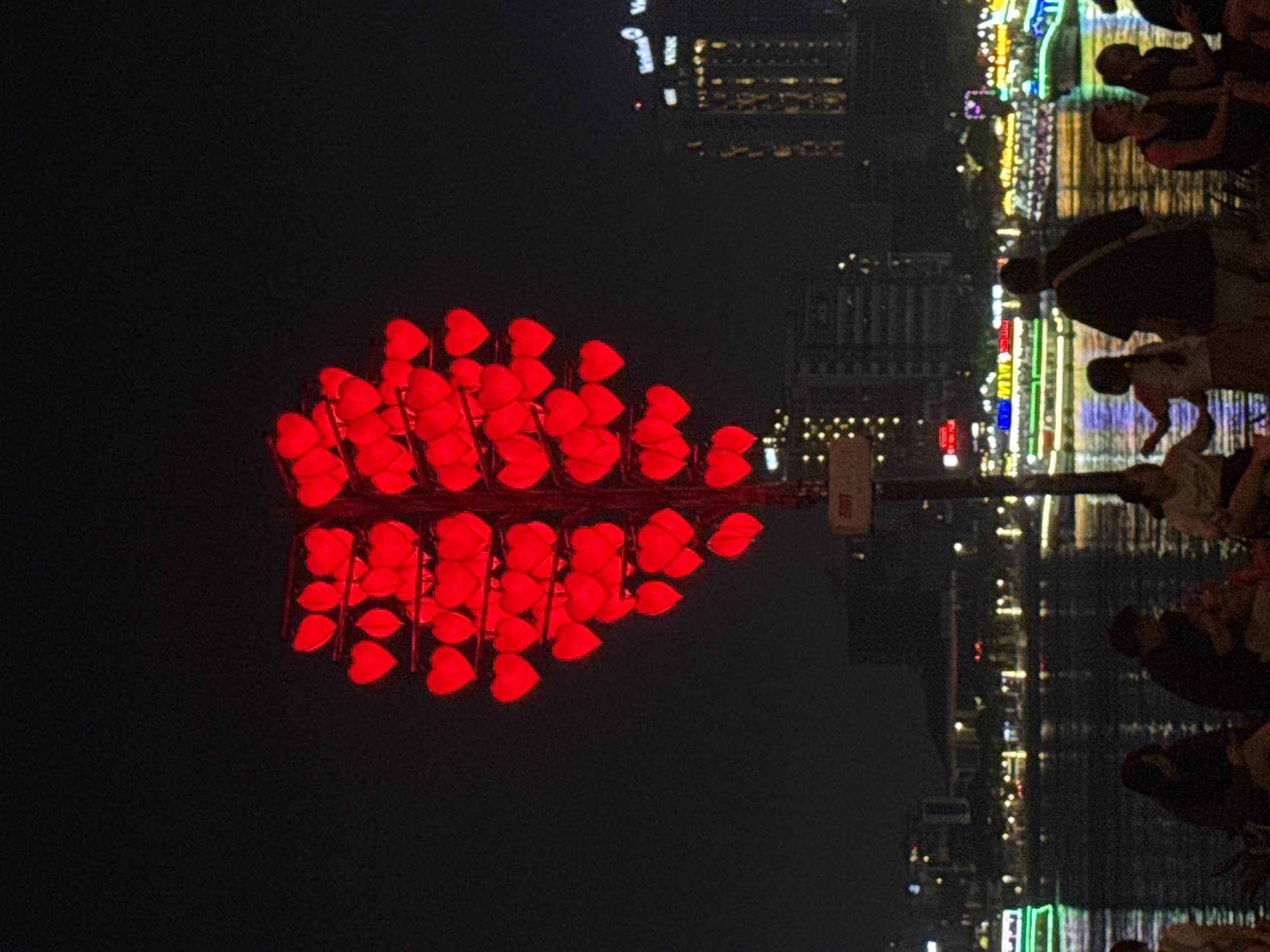 At night, the Hàn River in Đà Nẵng, Vietnam, is bustling with activity as people stroll along the waterfront. Lights twinkle from the city's buildings and reflect off the water's surface. In the foreground, a large, heart-shaped display made of red lights stands prominently, presumably to celebrate the love. People are present on the riverbank, engaging in conversations and enjoying the vibrant urban atmosphere.