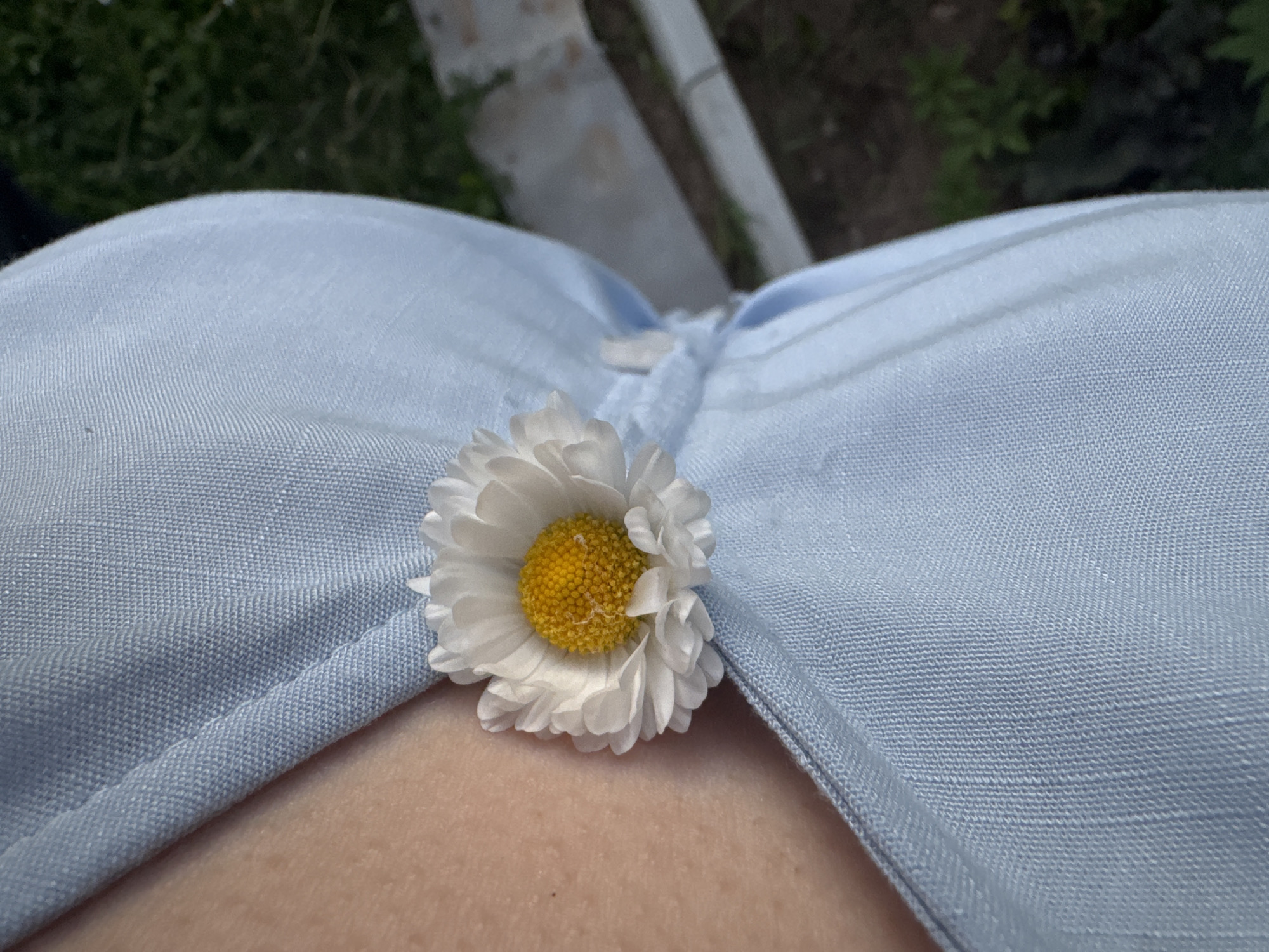A close-up, slightly angled overhead shot depicts a white daisy with a yellow center resting on a light blue collared garment, likely a shirt. The fabric of the garment shows a textured weave. Below the shirt, skin is visible. The upper portion of the image shows out-of-focus greenery and concrete elements, suggesting an outdoor setting. There is no information given to determine location.