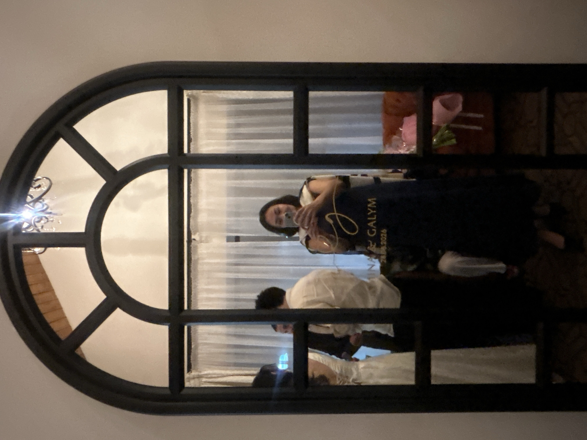 A woman is taking a selfie in a large, arched mirror, reflecting a scene inside a room. She is wearing a dark blue dress with gold lettering that reads "MINA & GALYM 28.08.2026." In the reflection, behind her, a bride in a white dress and a groom in a light-colored shirt are visible, suggesting a wedding event. To the right of the mirror, a section of an orange sofa and two pink gift bags are visible. The room appears to be decorated with sheer white curtains and a chandelier, hinting at a formal or celebratory setting. The image does not provide enough visual information to confirm a specific location in Almaty, Kazakhstan.
