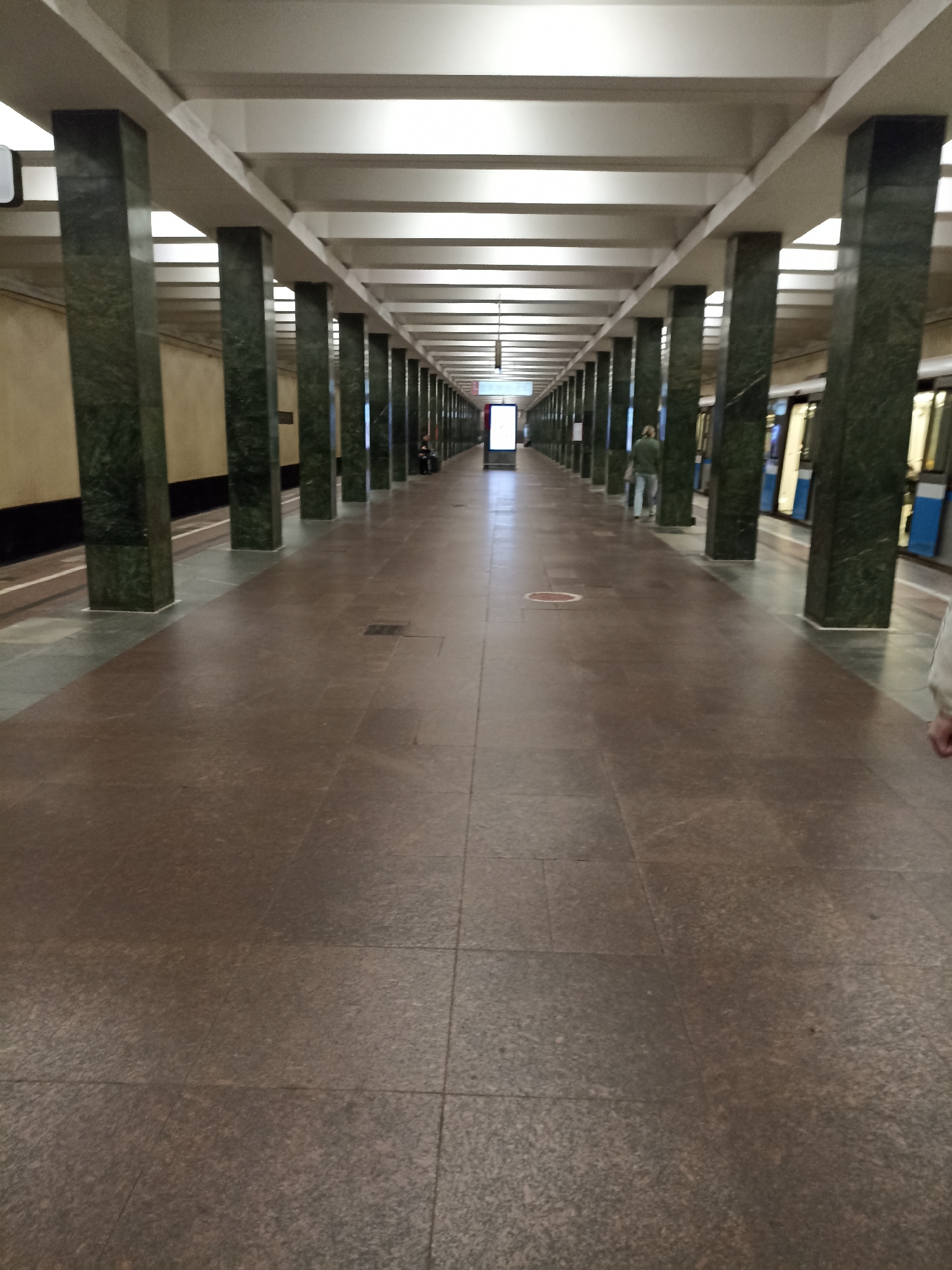 This is a vertical, eye-level, full-shot photograph taken inside a Moscow Metro station. The perspective is looking down a long, symmetrical platform, with a train visible on the right side of the image. The station features a repeating pattern of dark green marble-clad pillars supporting a white, beamed ceiling. The platform floor is tiled in a reddish-brown granite. In the background, a few people are visible, including one person standing on the platform and another walking away from the camera. Further down the platform, a lit advertisement screen is visible, and at the very end, the tunnel opening is apparent. The overall impression is one of depth and order, characteristic of many Moscow Metro stations.