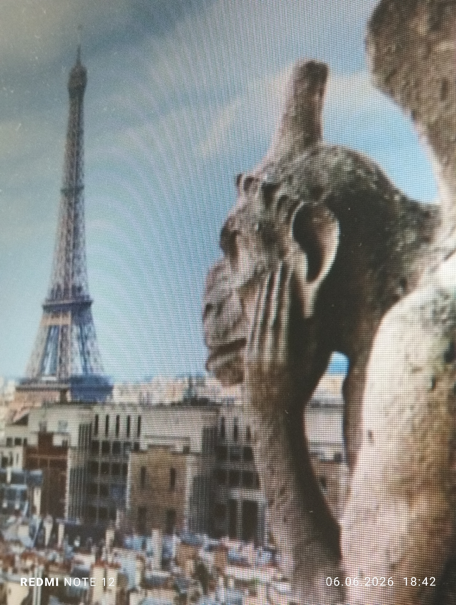A digitally captured image, displaying a visible pixel grid, shows a photograph of a screen. The content on the screen depicts a panoramic view of Paris, France, from an elevated vantage point. In the immediate foreground, to the right, a weathered stone gargoyle, likely from Notre Dame Cathedral, is partially visible and slightly out of focus, with its right ear and parts of its head clearly discernible. The middle ground reveals a dense cityscape of Parisian buildings with various architectural styles, including rooftops and facades. Dominating the background on the left is the iconic Eiffel Tower, its intricate lattice structure clearly visible against a light blue sky with faint cloud cover. The overall scene on the screen is bright and rendered in natural colors. Watermarks are present on the photograph of the screen: 'REDMI NOTE 12' is visible in white text in the bottom left corner, and '06.06.2026 18:42' is visible in white text in the bottom right corner, indicating the device used and the date/time of capture. This image was captured in the city of Burgas, Bulgaria.